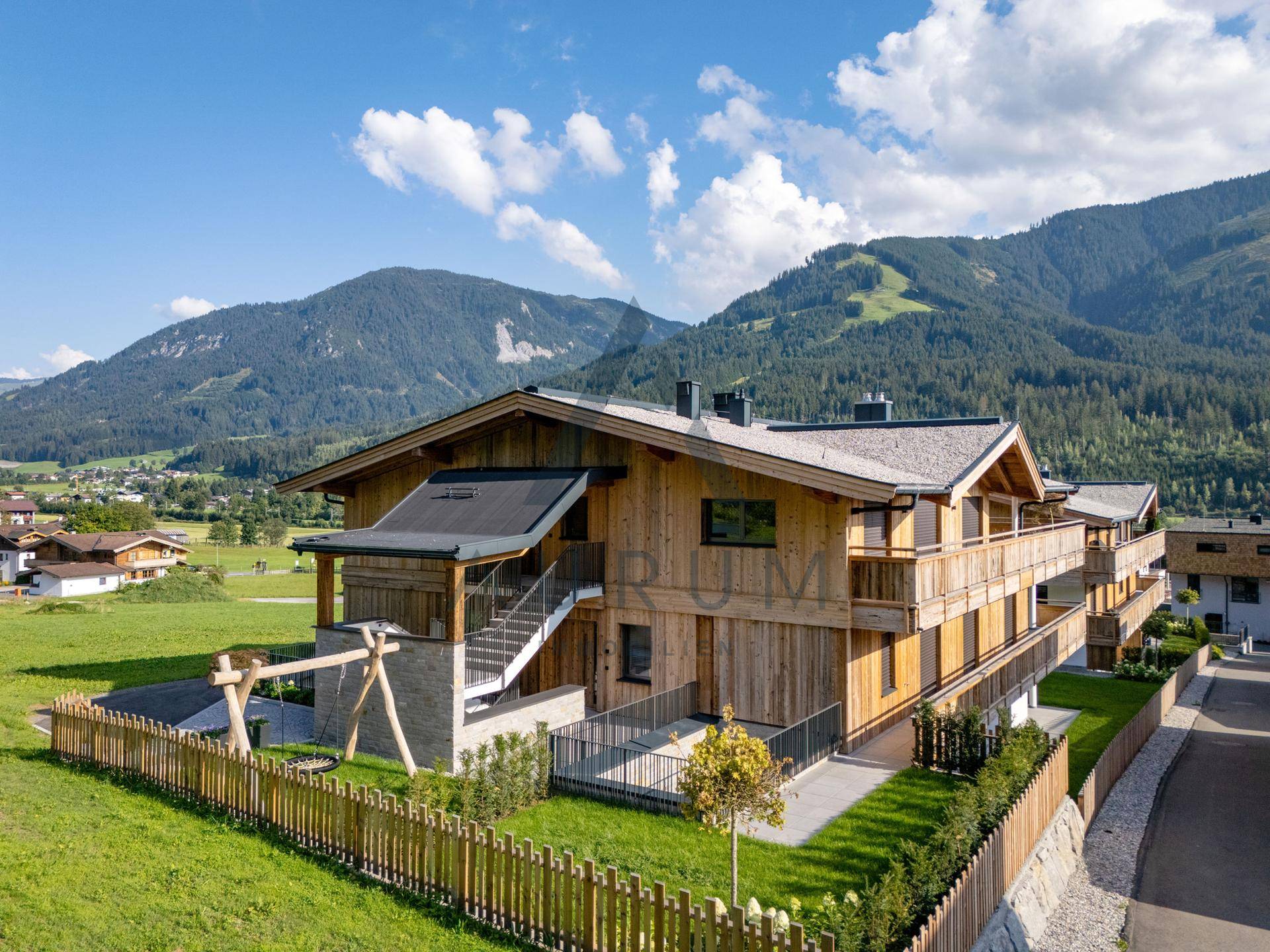 Seitlicher Blick auf das Holzhaus mit eingezäuntem Garten und Kinderschaukel, umgeben von Grün.