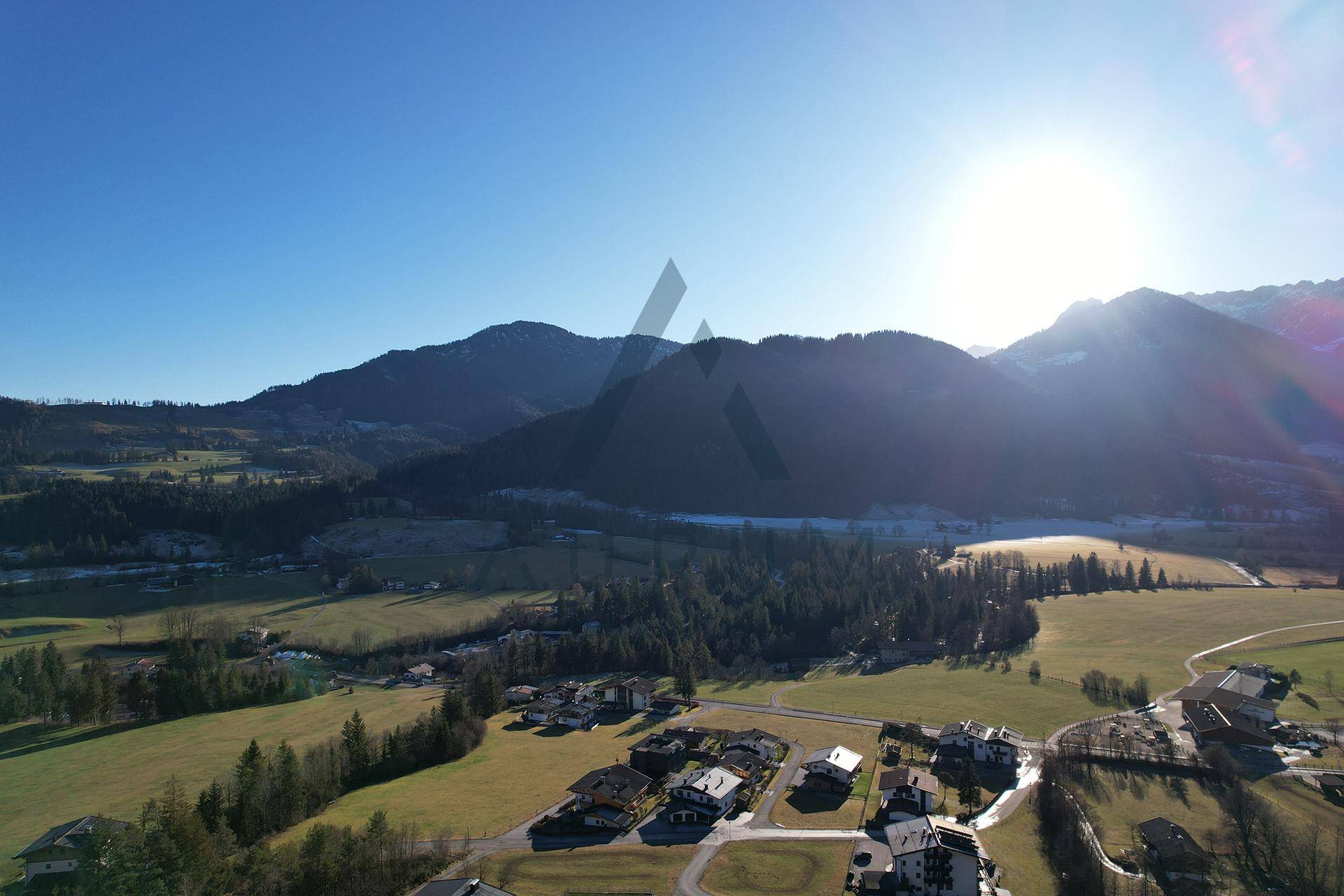 Panoramische Luftaufnahme eines sonnigen Tals mit Bergen, weiten Feldern und einer Siedlung, beleuchtet von der tiefstehenden Sonne.