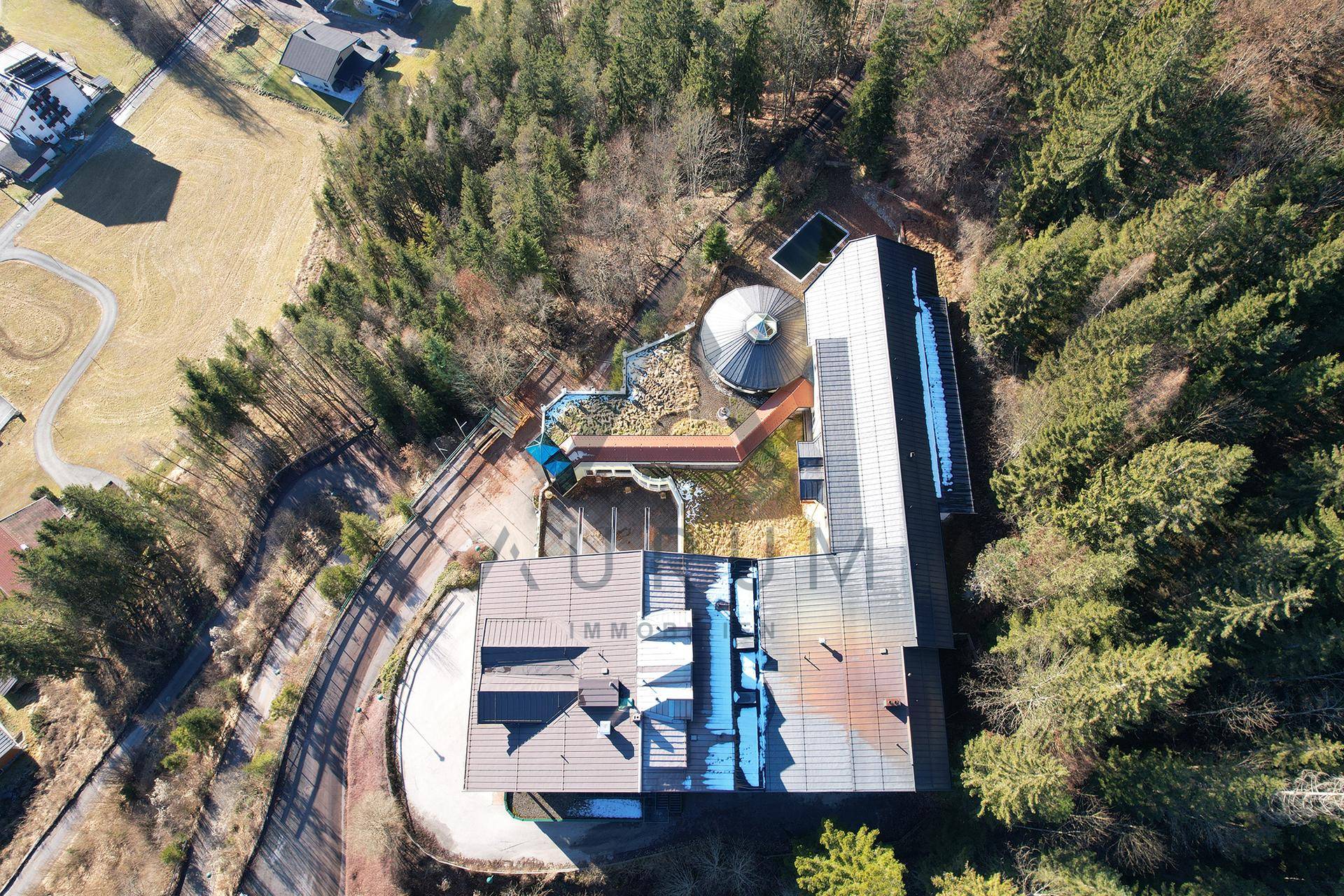 Senkrechte Luftaufnahme des Gebäudekomplexes, umgeben von Wald, mit sichtbaren Dachstrukturen und Außenbereichen.