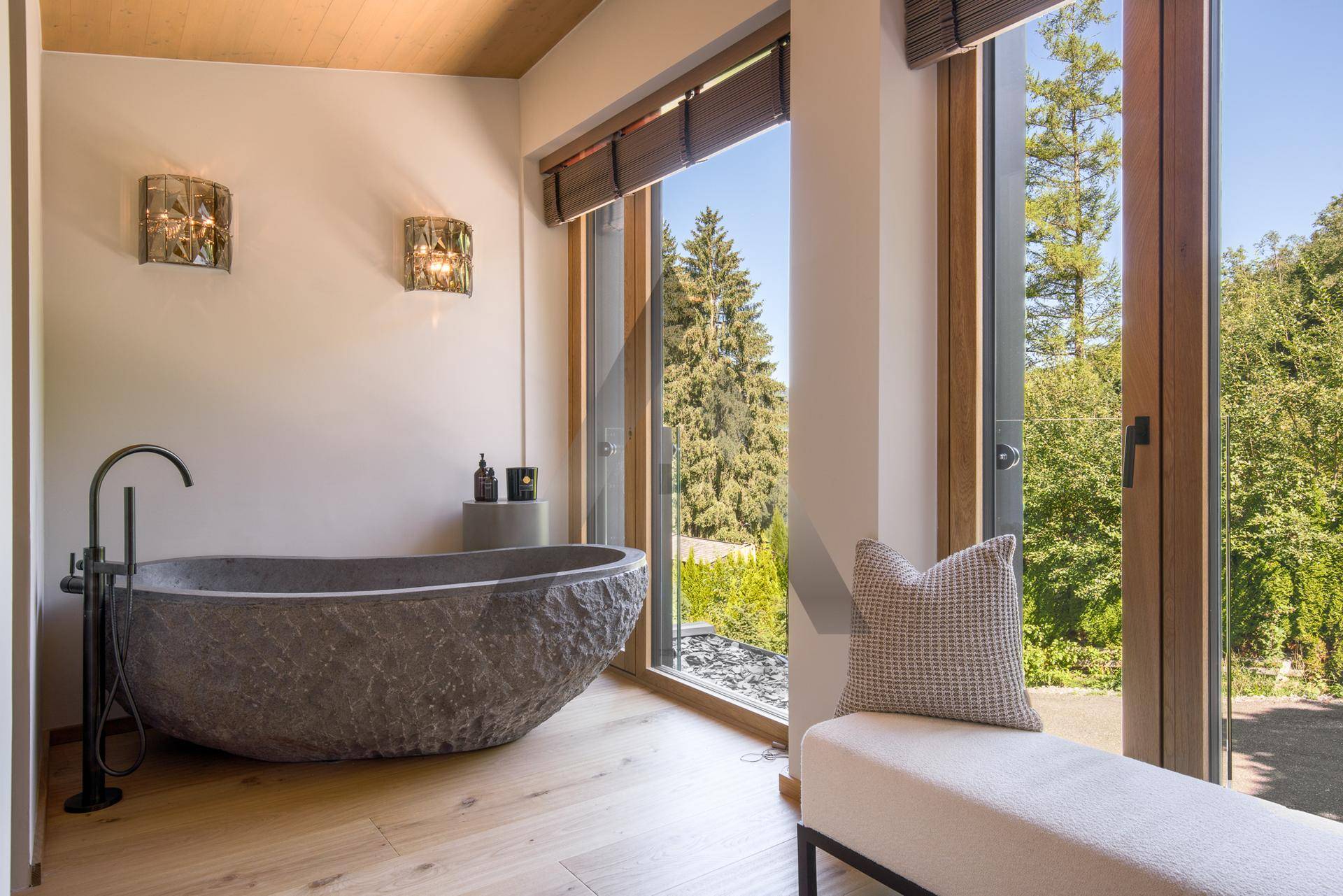 Luxuriöses Badezimmer mit freistehender Steinbadewanne und großen Fenstern mit Waldblick.