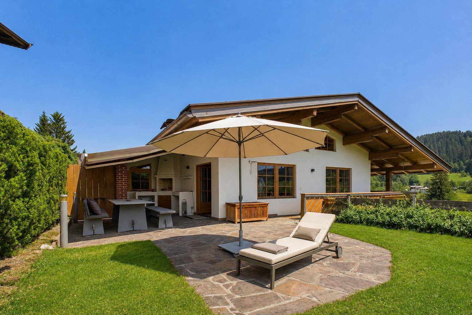 Sonniger Terrassenbereich mit Liegestuhl, Sonnenschirm und Grillplatz im Garten.