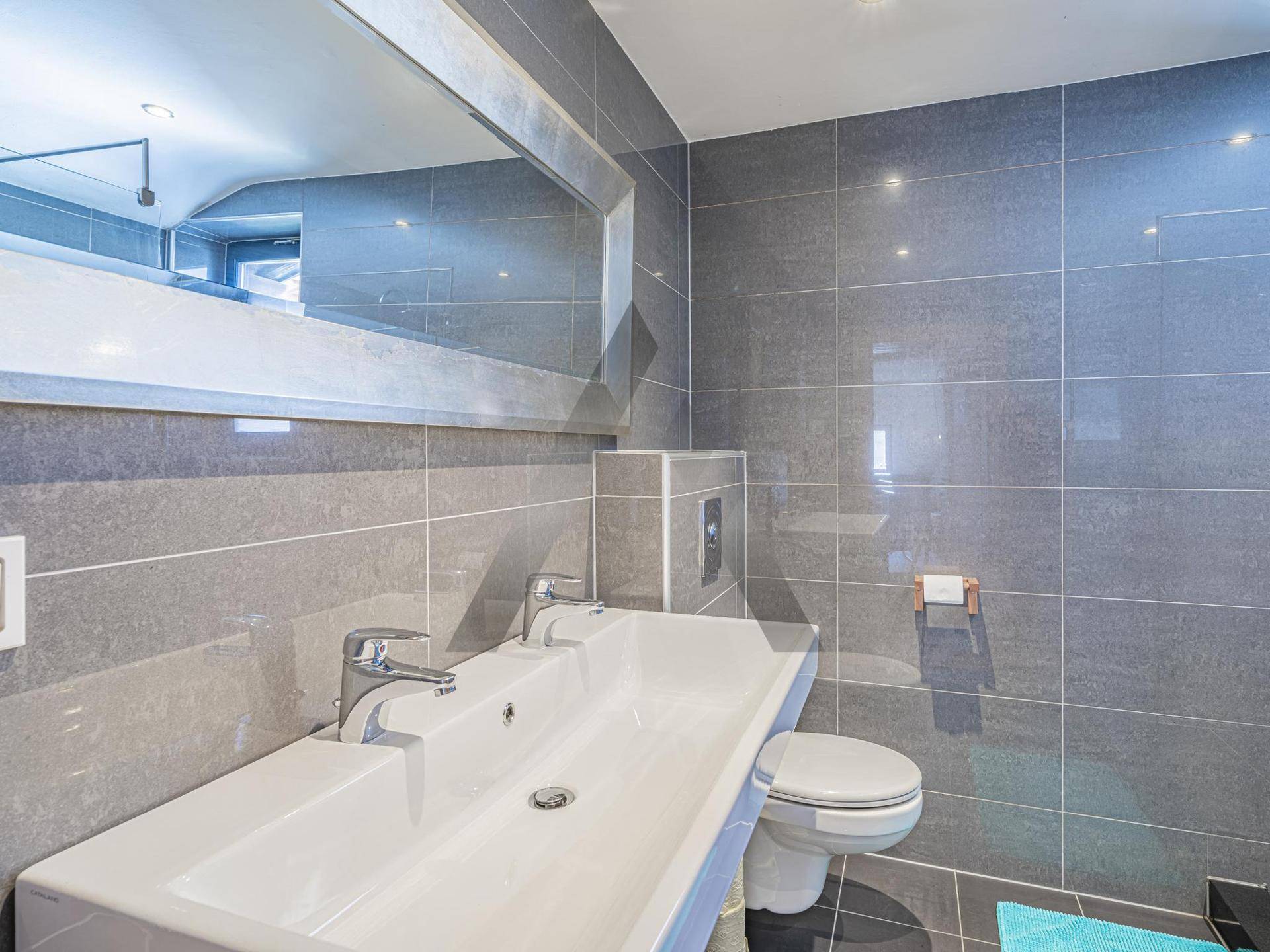 Modernes Badezimmer mit grauen Fliesen, Doppelwaschbecken und wandmontierter Toilette.
