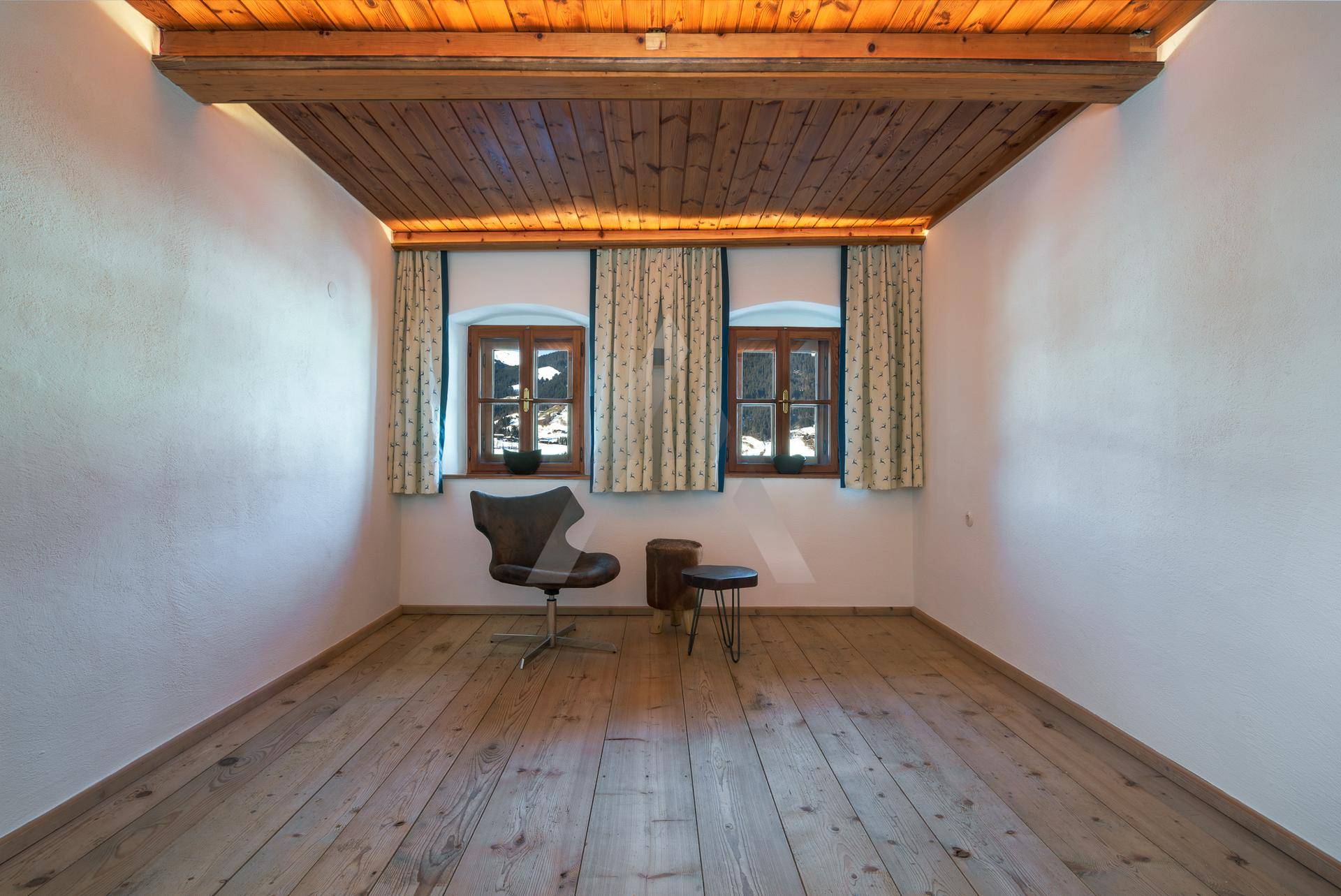 Helles Zimmer mit Holzboden, Holzbalkendecke und zwei Fenstern.