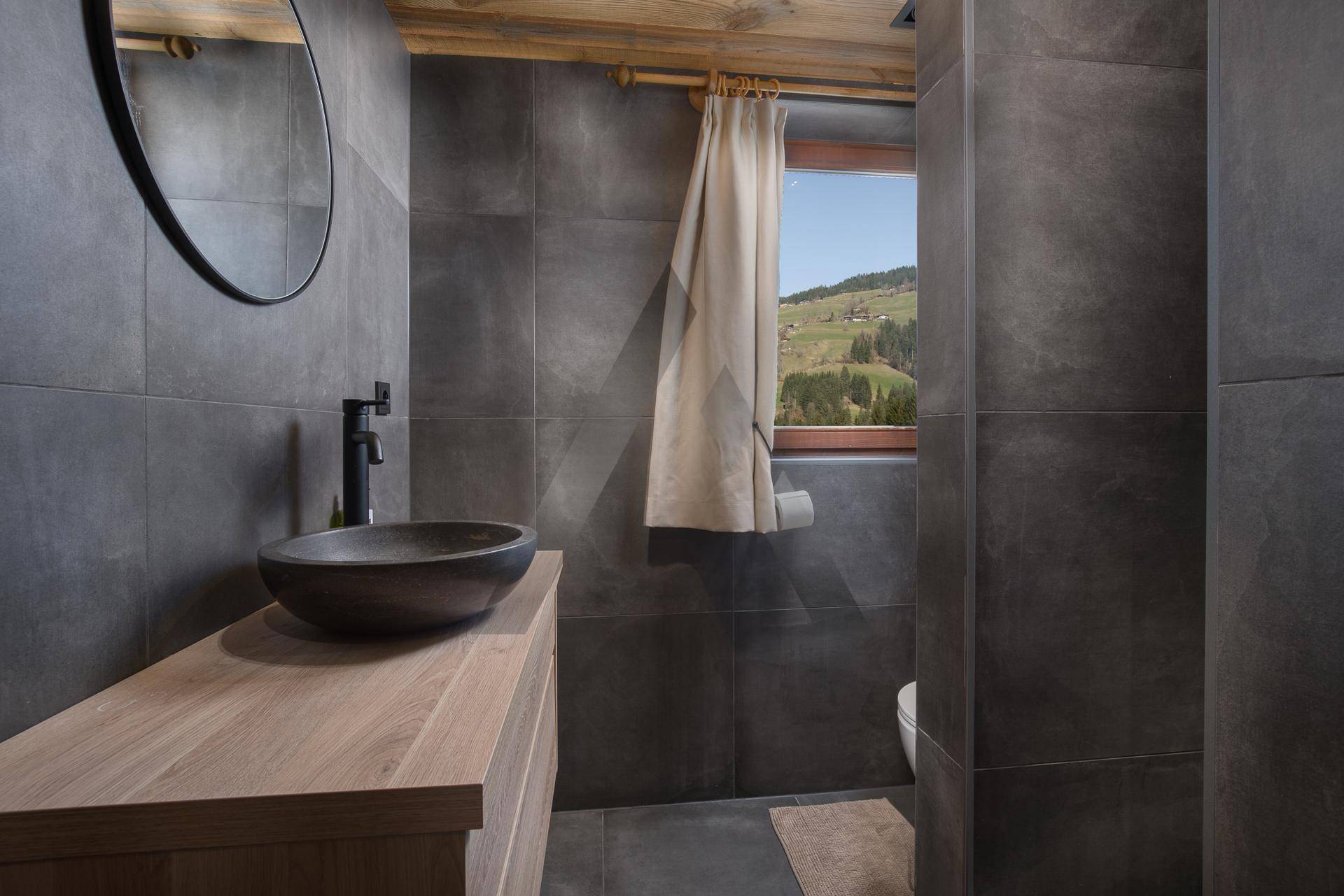 Modernes Badezimmer mit dunklen Fliesen, Holzwaschtisch und Fenster mit Bergblick.