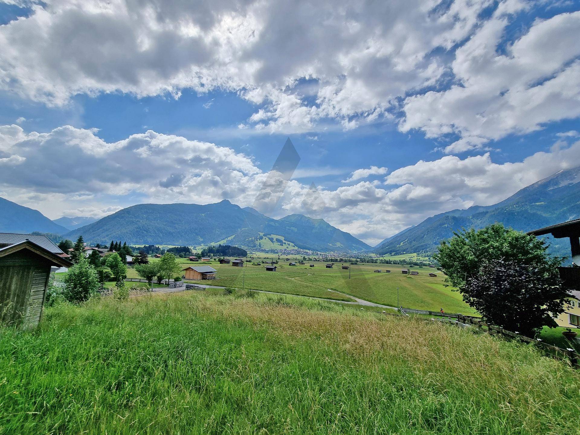 Panoramablick über weite grüne Felder und die majestätische Bergkette unter einem blauen Himmel mit Wolken.