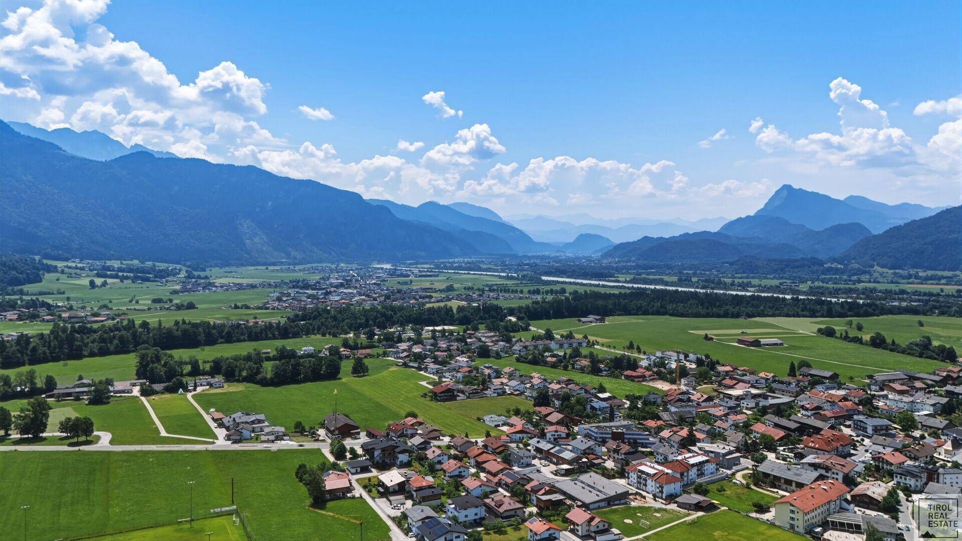 Panoramablick auf ein malerisches Dorf im Tal, umgeben von grünen Feldern und majestätischen Bergen unter blauem Himmel.