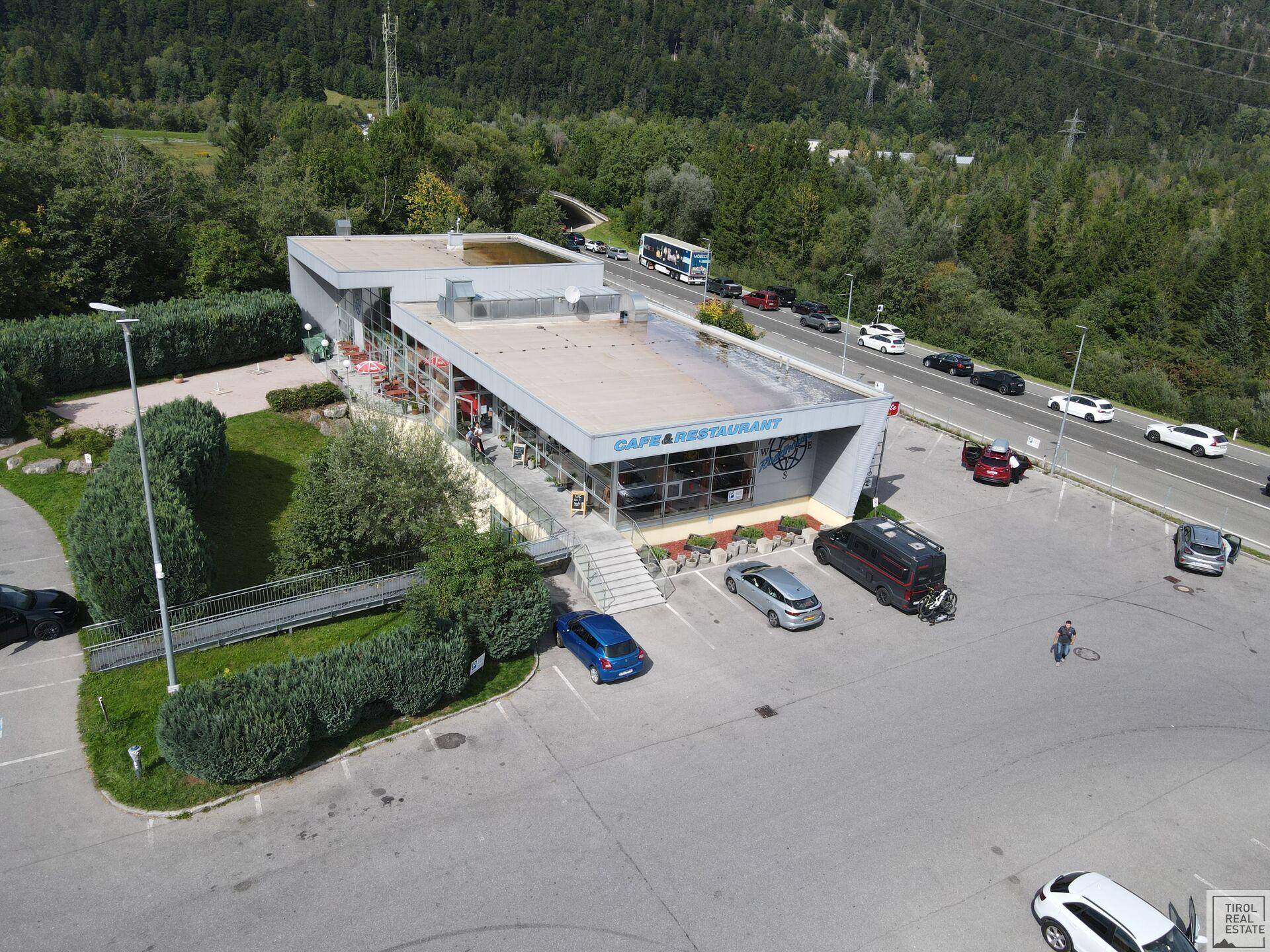 Luftaufnahme des modernen Gebäudes mit großem Parkplatz und umliegender Berglandschaft.