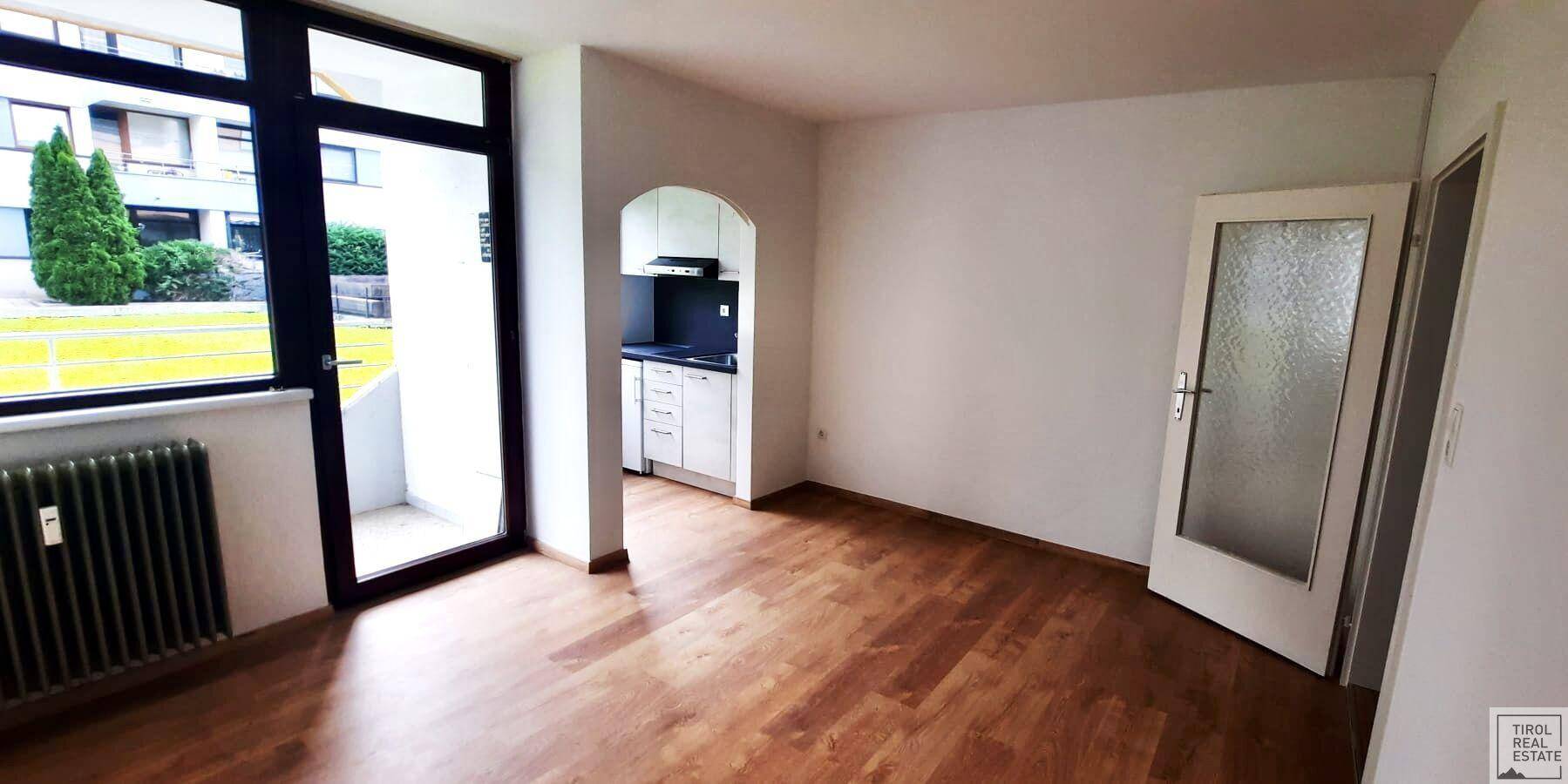 Leerer Wohnraum mit Holzboden, großem Fenster und integrierter Küchennische.