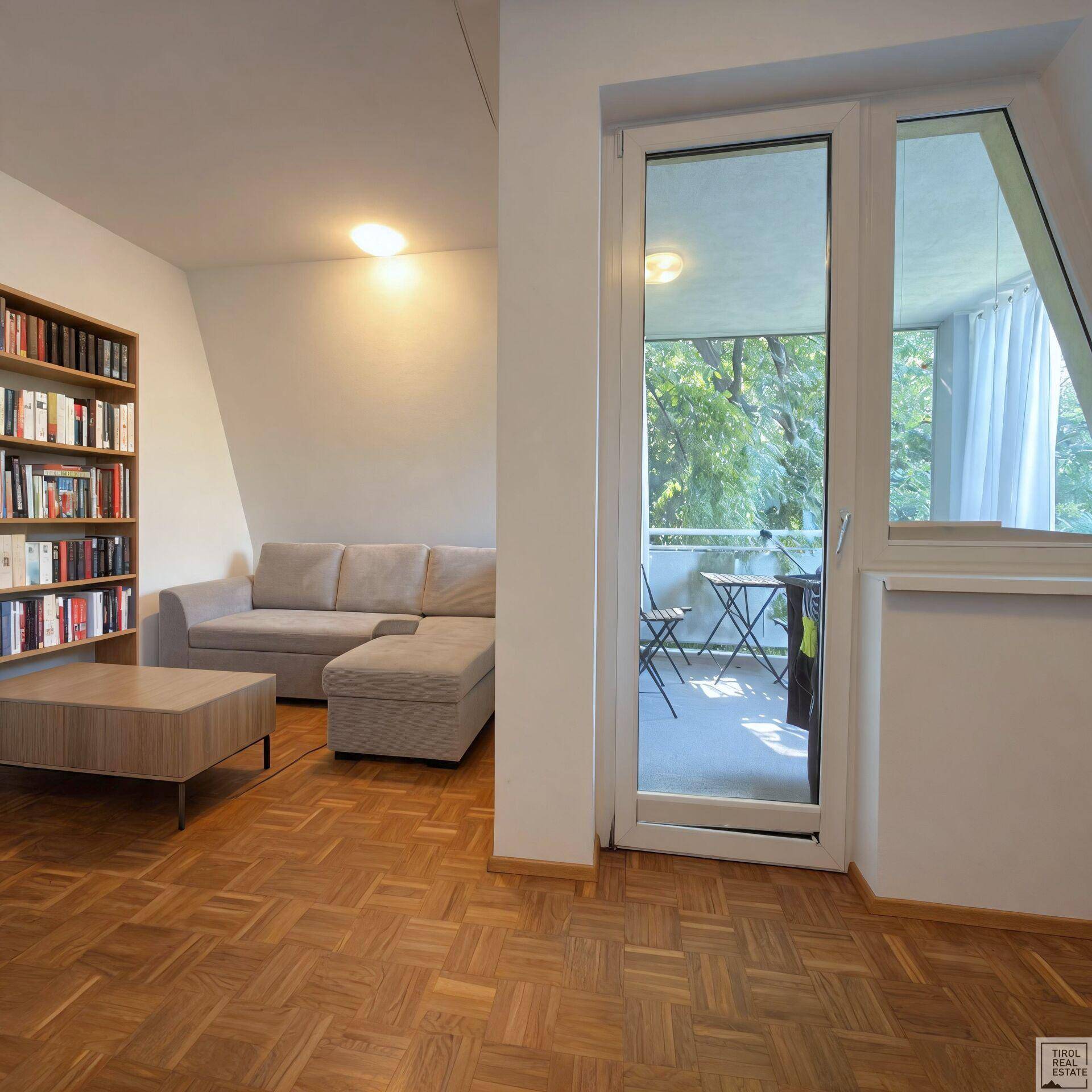 Wohnbereich mit Sofa, Parkettboden und Zugang zu einem Balkon, der eine grüne Aussicht bietet.