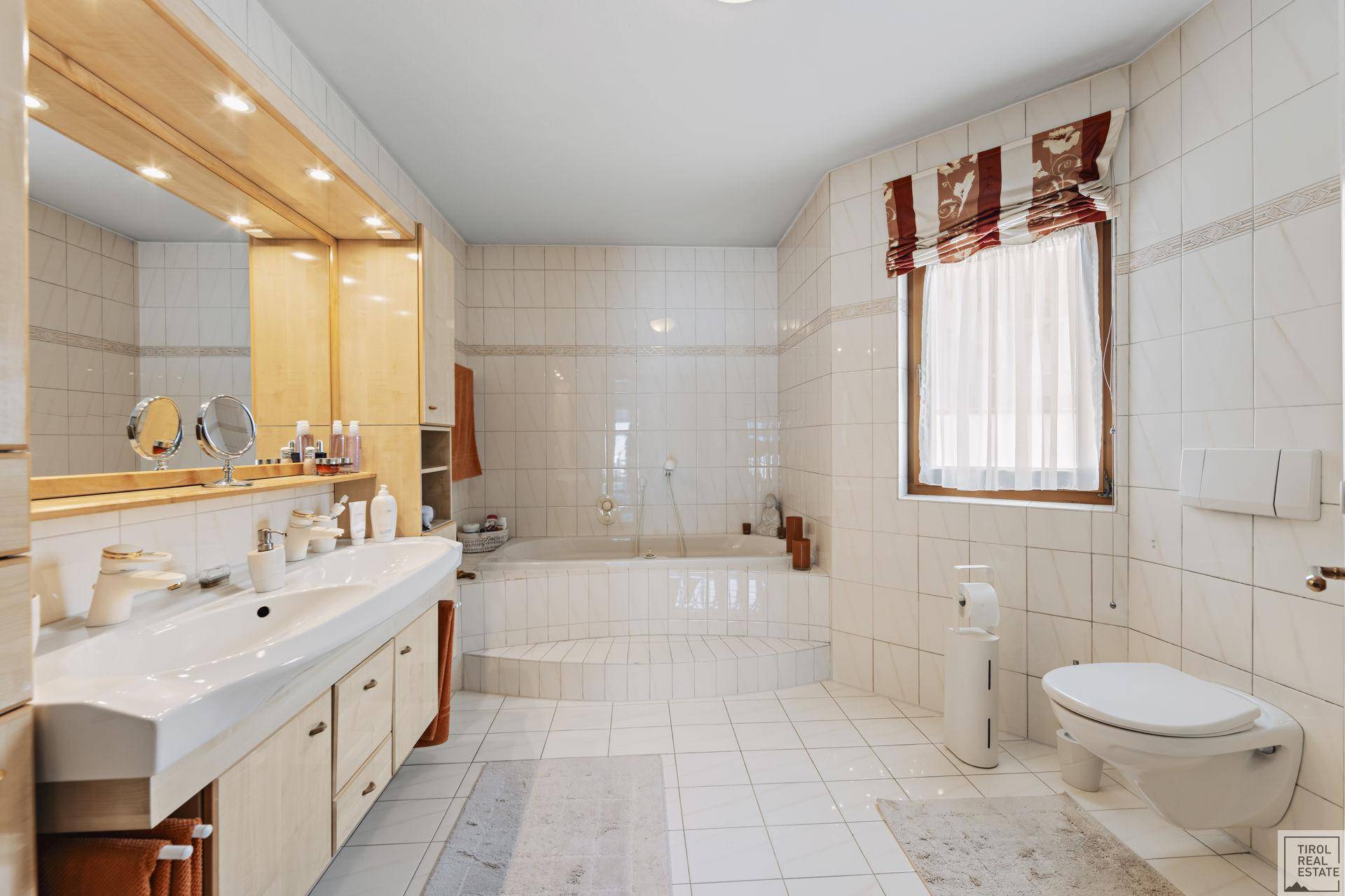 Badezimmer mit weißen Fliesen, Toilette, Doppelwaschbecken und einem Fenster für Tageslicht.