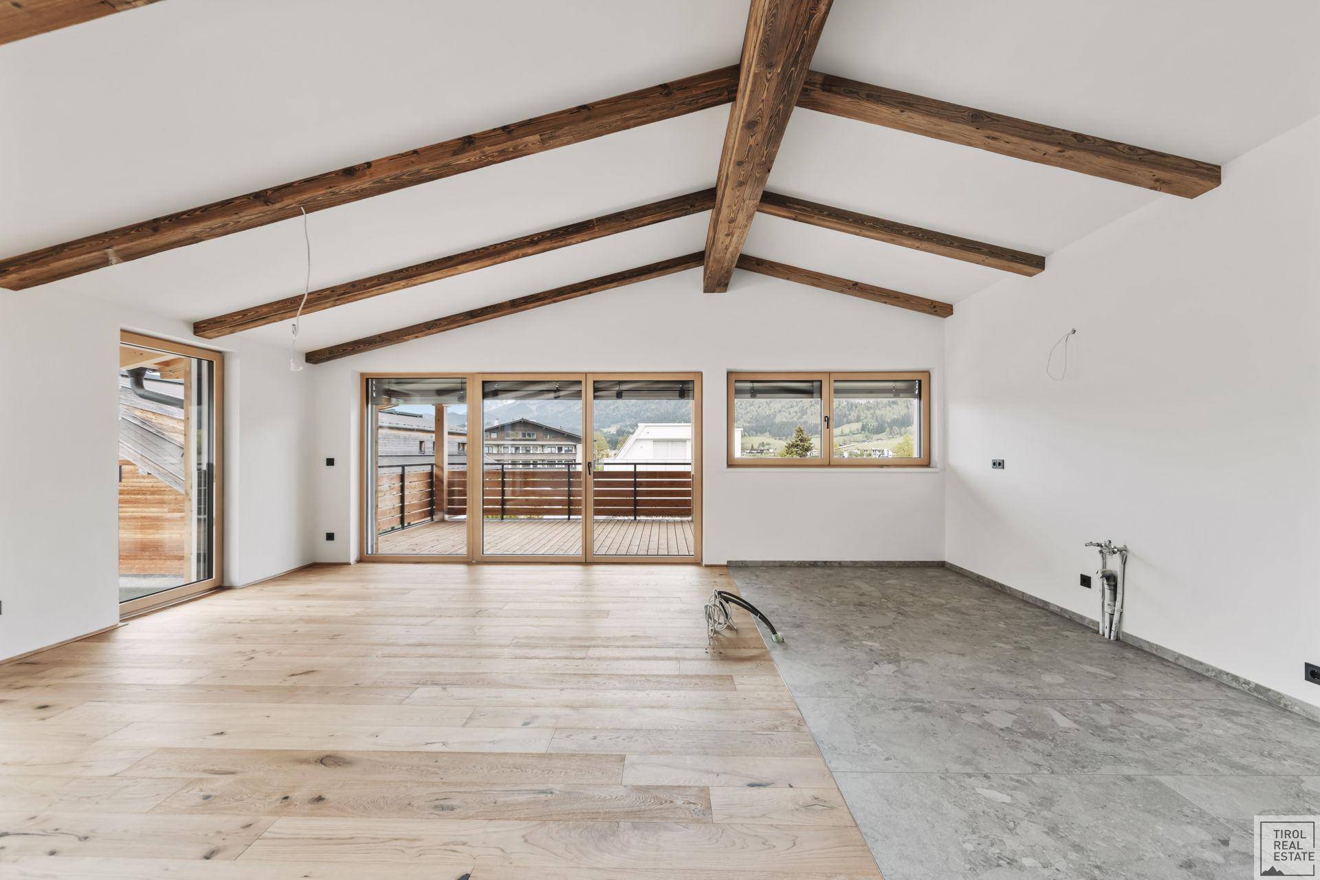 Heller, unmöblierter Raum mit Holzbalken, Parkettboden und breiter Fensterfront zur Terrasse.