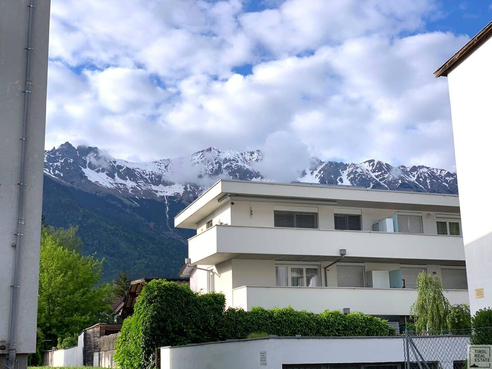 Moderne Wohnanlage mit Balkonen und gepflegten Hecken vor der beeindruckenden Kulisse schneebedeckter Berge.