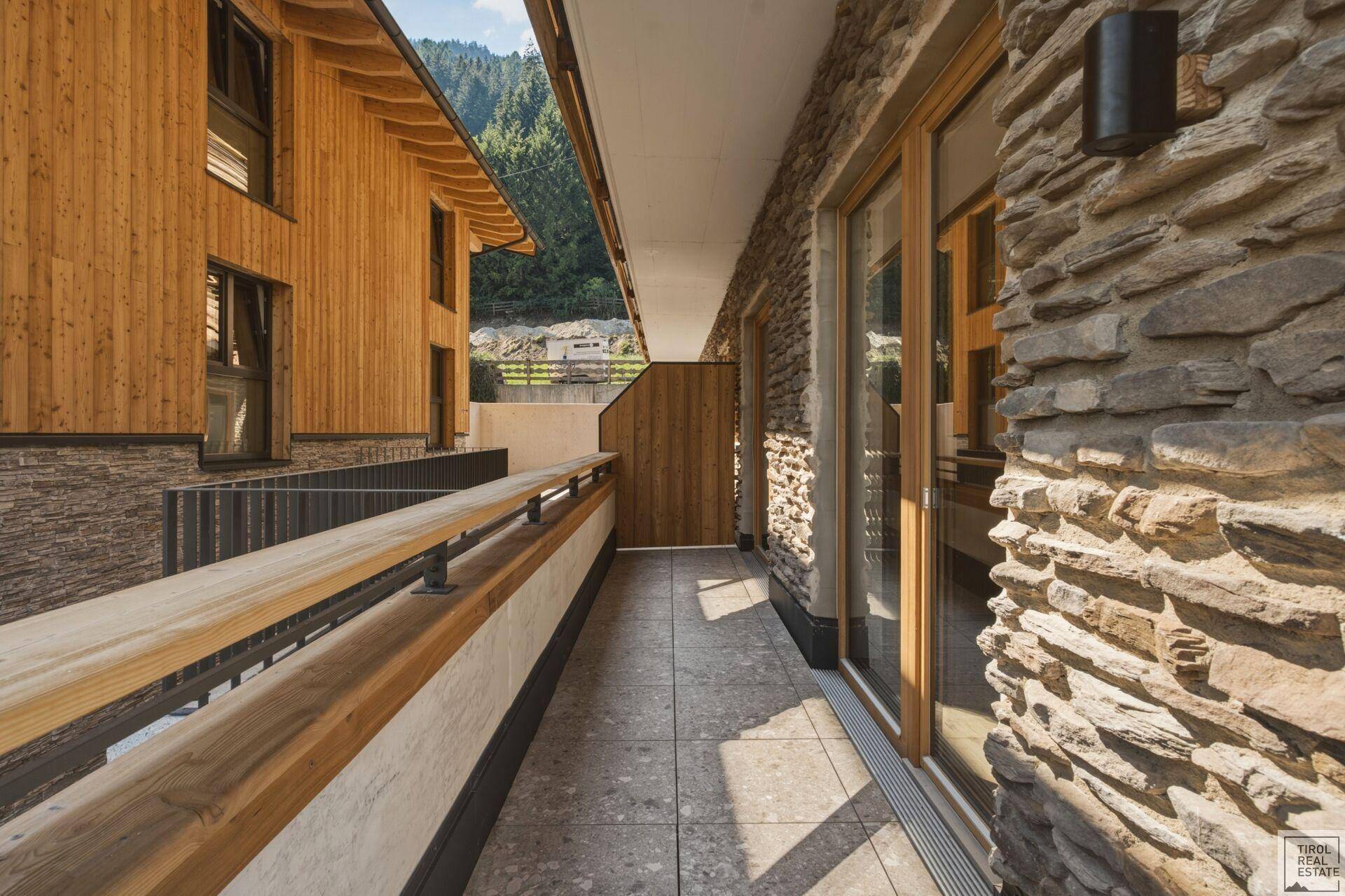 Langer Balkon mit Steinwand und Holzelementen, umgeben von alpiner Natur.