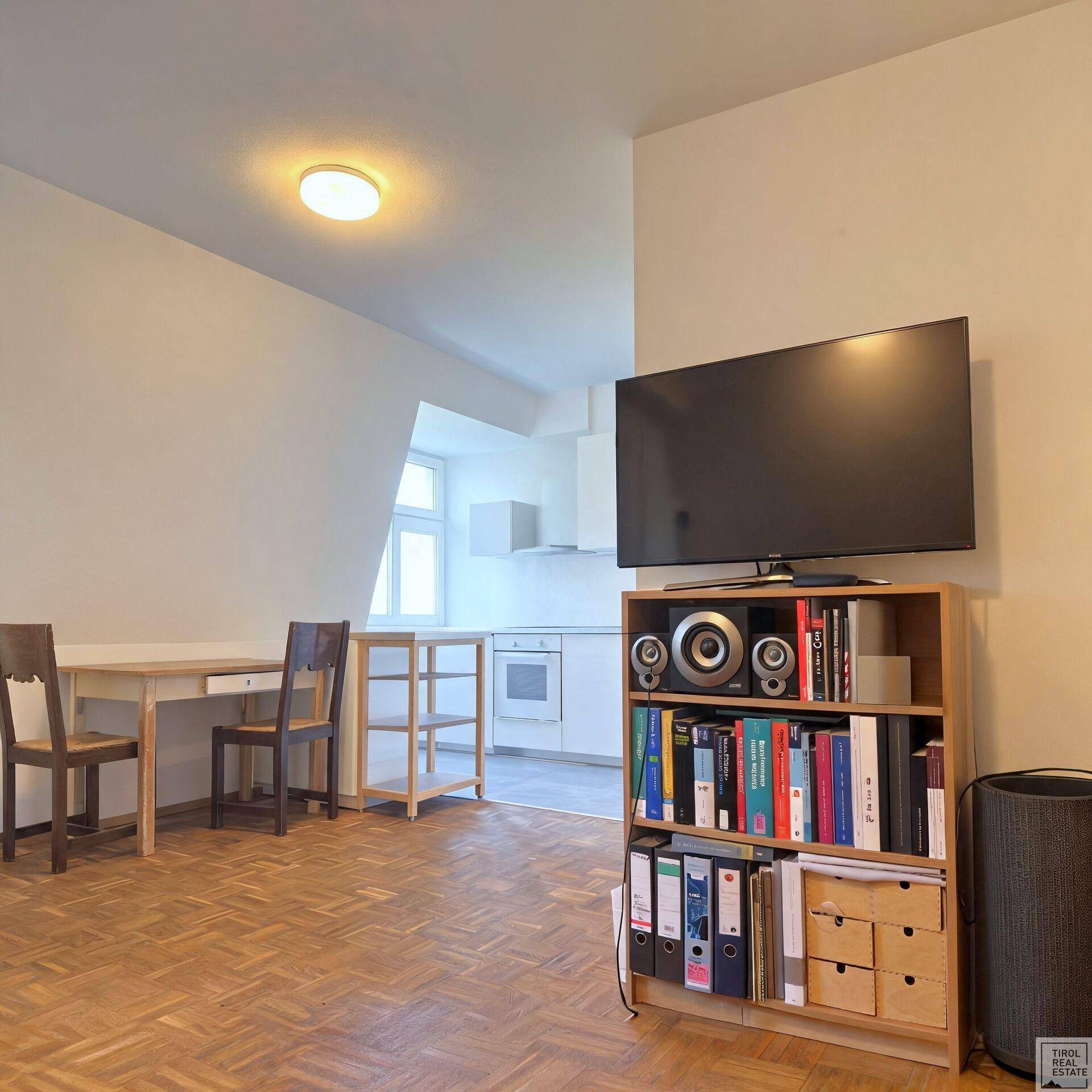 Offener Wohn- und Essbereich mit Fernseher, Bücherregal und integrierter Küche im hinteren Bereich.