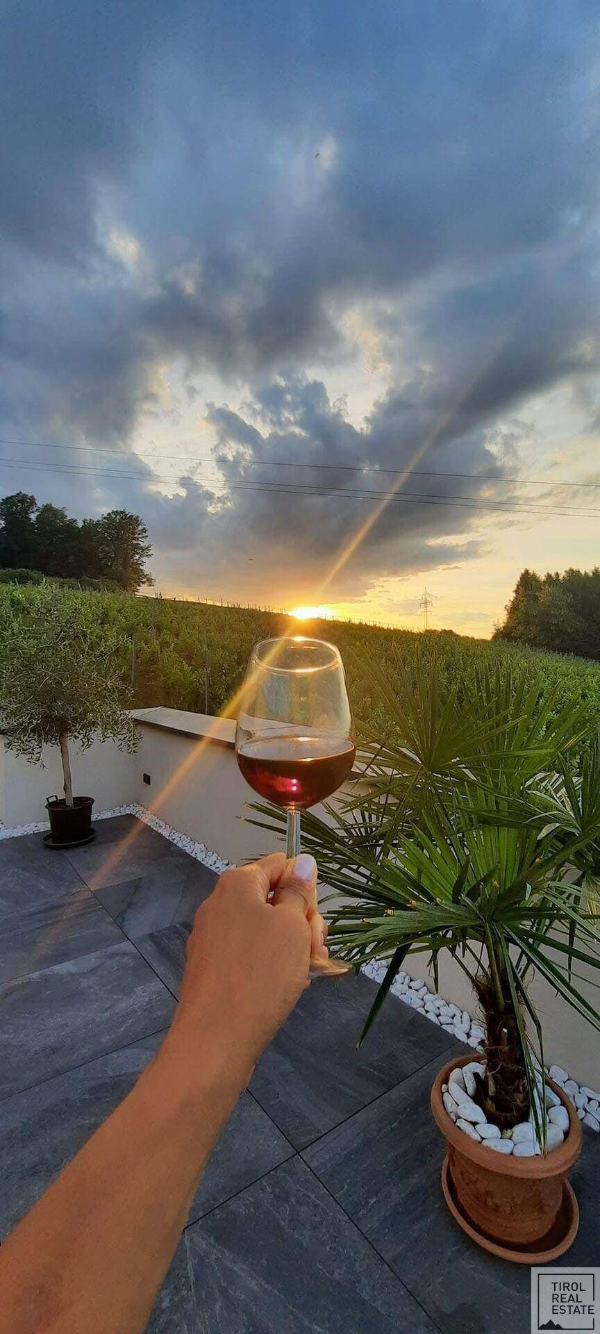 Hand hält ein Weinglas auf einer Terrasse mit Blick auf einen Sonnenuntergang über Weinbergen.