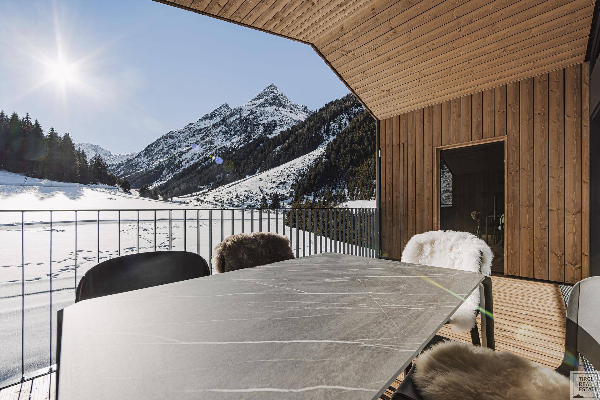 Sonniger Balkon mit Esstisch und Stühlen, umgeben von einer beeindruckenden verschneiten Berglandschaft.