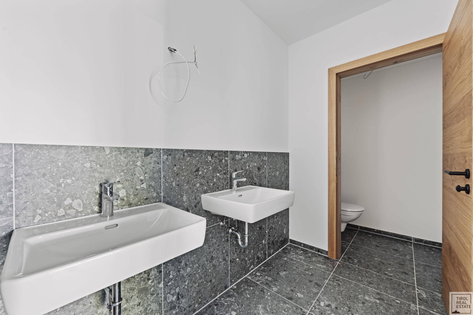 Modernes Badezimmer mit zwei Waschbecken und separatem WC-Bereich, ausgestattet mit grauen Fliesen.