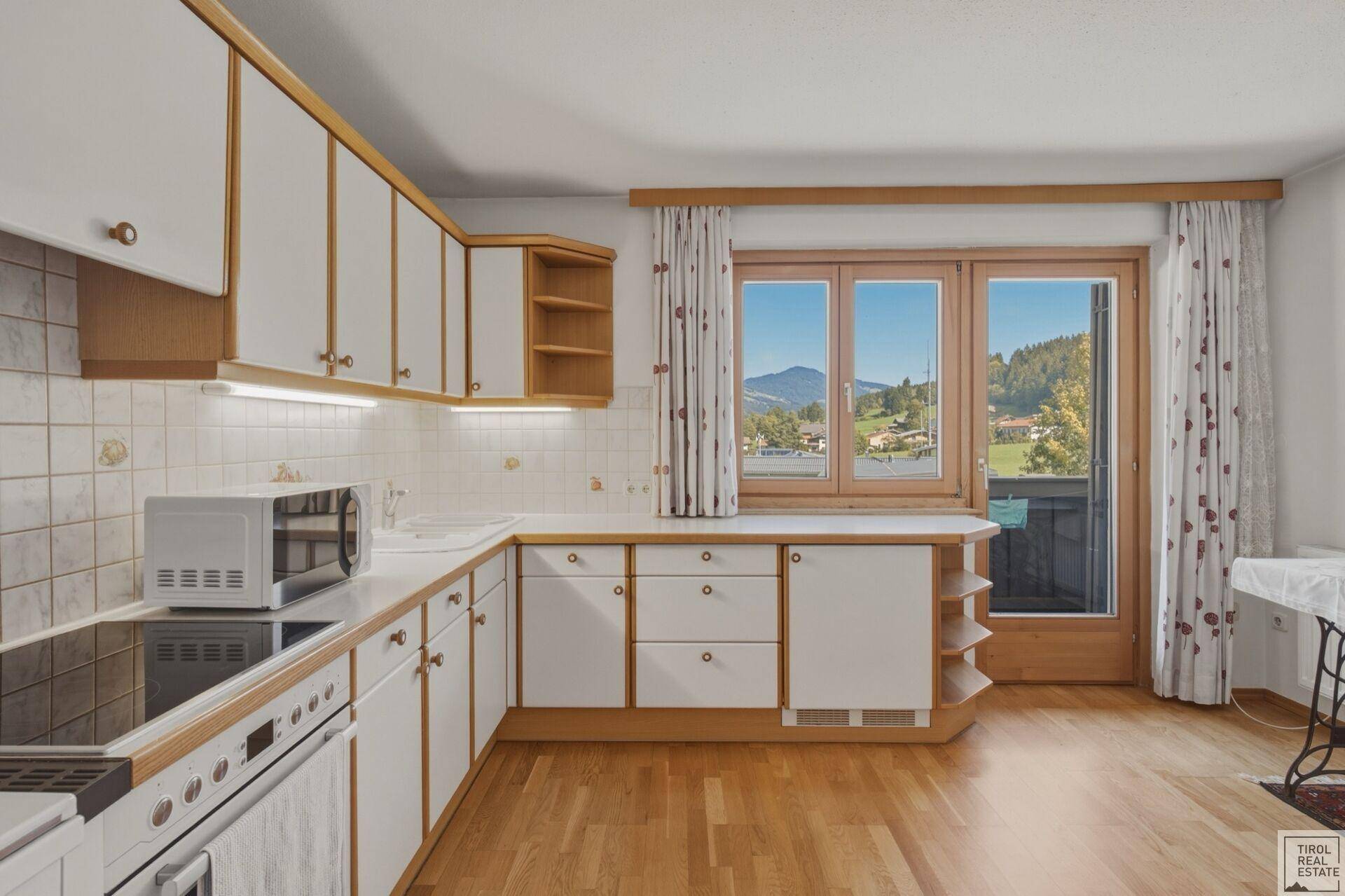 Helle Küche mit Holzboden, weißen Fronten und Zugang zu einem Balkon mit Bergblick.