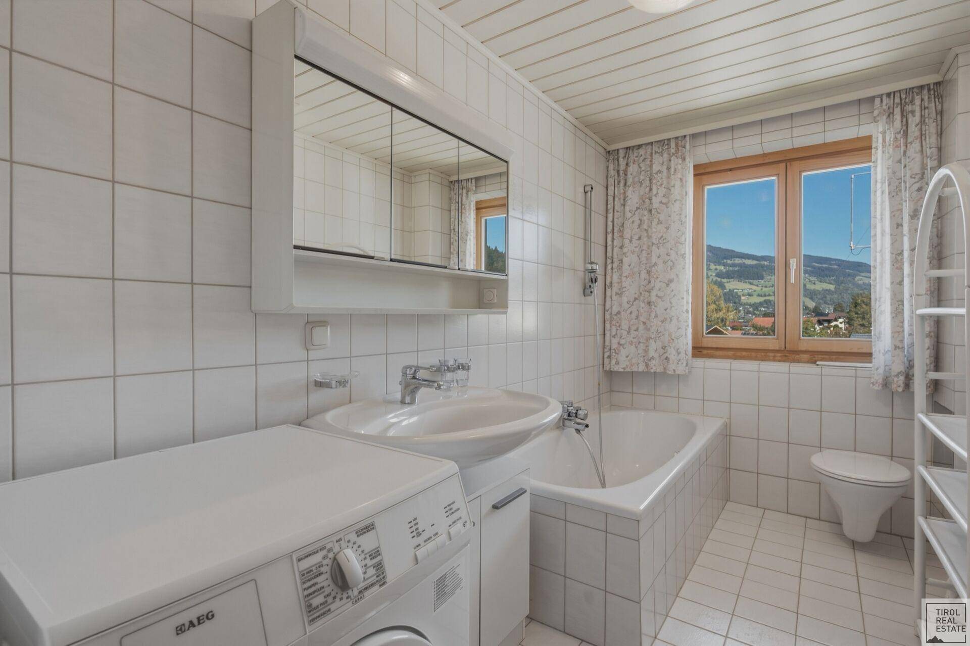 Helles Badezimmer mit weißen Fliesen, Badewanne und Waschmaschine unter dem Waschbecken.