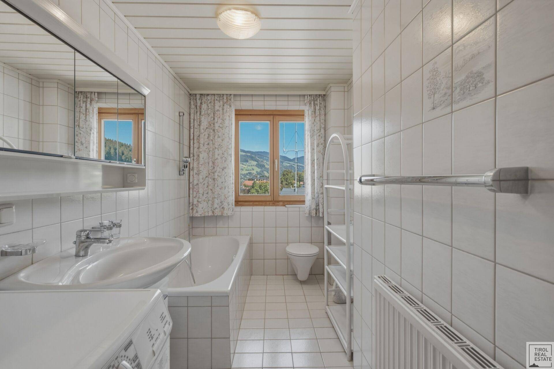 Gefliestes Badezimmer mit Badewanne, Waschbecken und Fenster mit Bergblick.