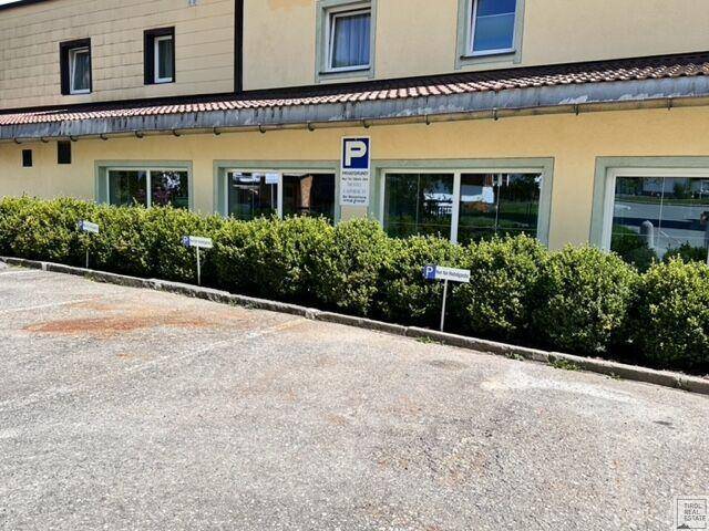 Gebäudefassade mit großen Fenstern und einem Parkplatz, abgegrenzt durch eine grüne Hecke.
