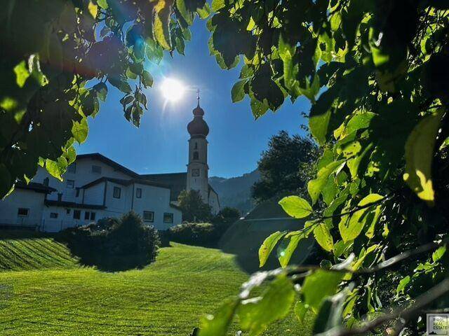 Idyllischer Blick auf eine Kirche und grüne Hügel, umrahmt von Blättern im Sonnenlicht.