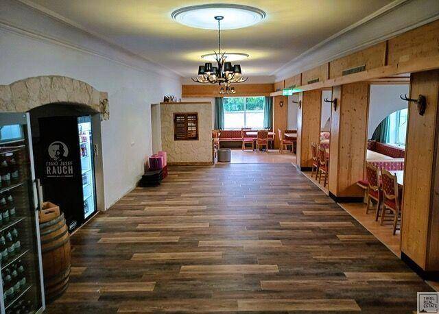 Geräumiger Gemeinschaftsraum mit Holzboden, Deckenleuchten und Essbereichen im Hintergrund.
