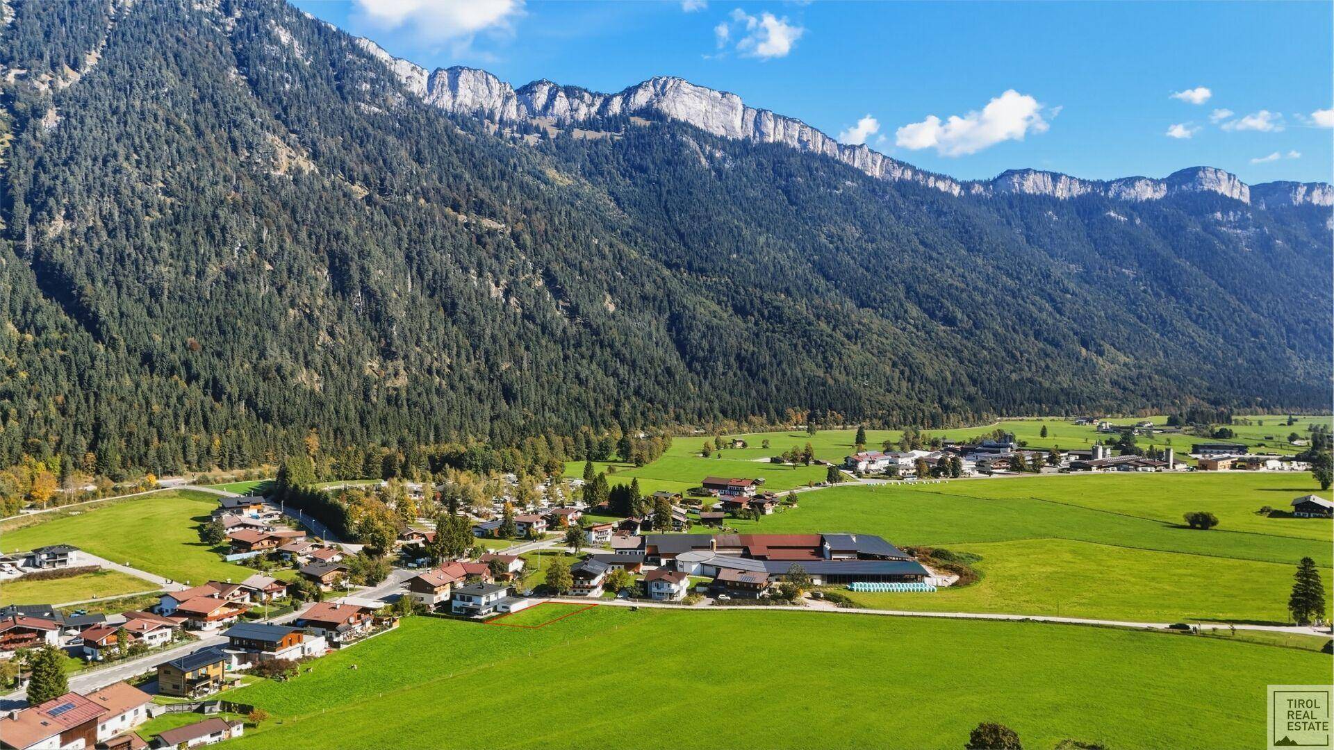 Malerische Luftaufnahme eines Dorfes in einem grünen Tal, umgeben von imposanten bewaldeten Bergen.