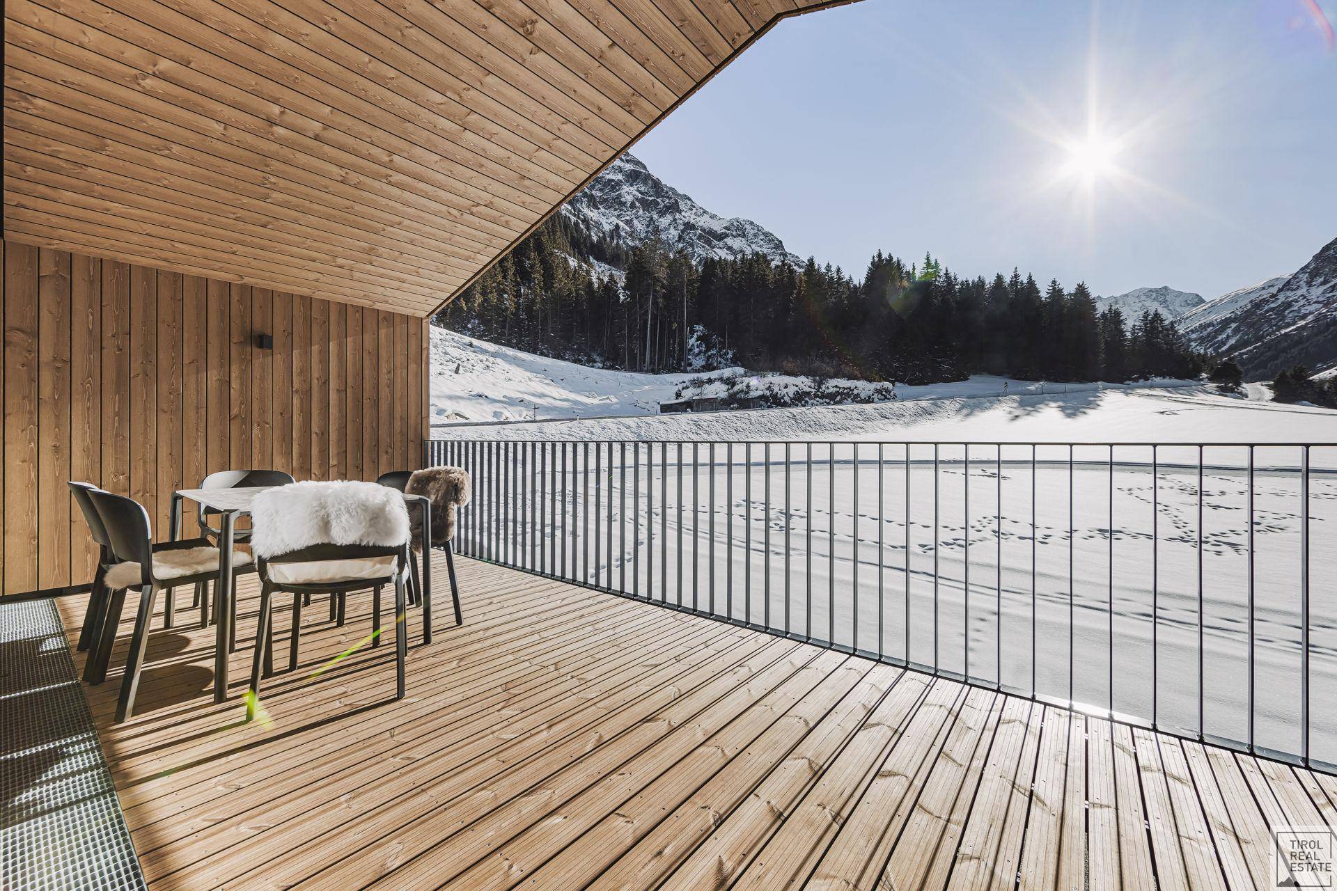 Geräumiger Holzbalkon mit Sitzgelegenheiten und weitem Blick auf die verschneite Bergwelt unter strahlendem Sonnenschein.