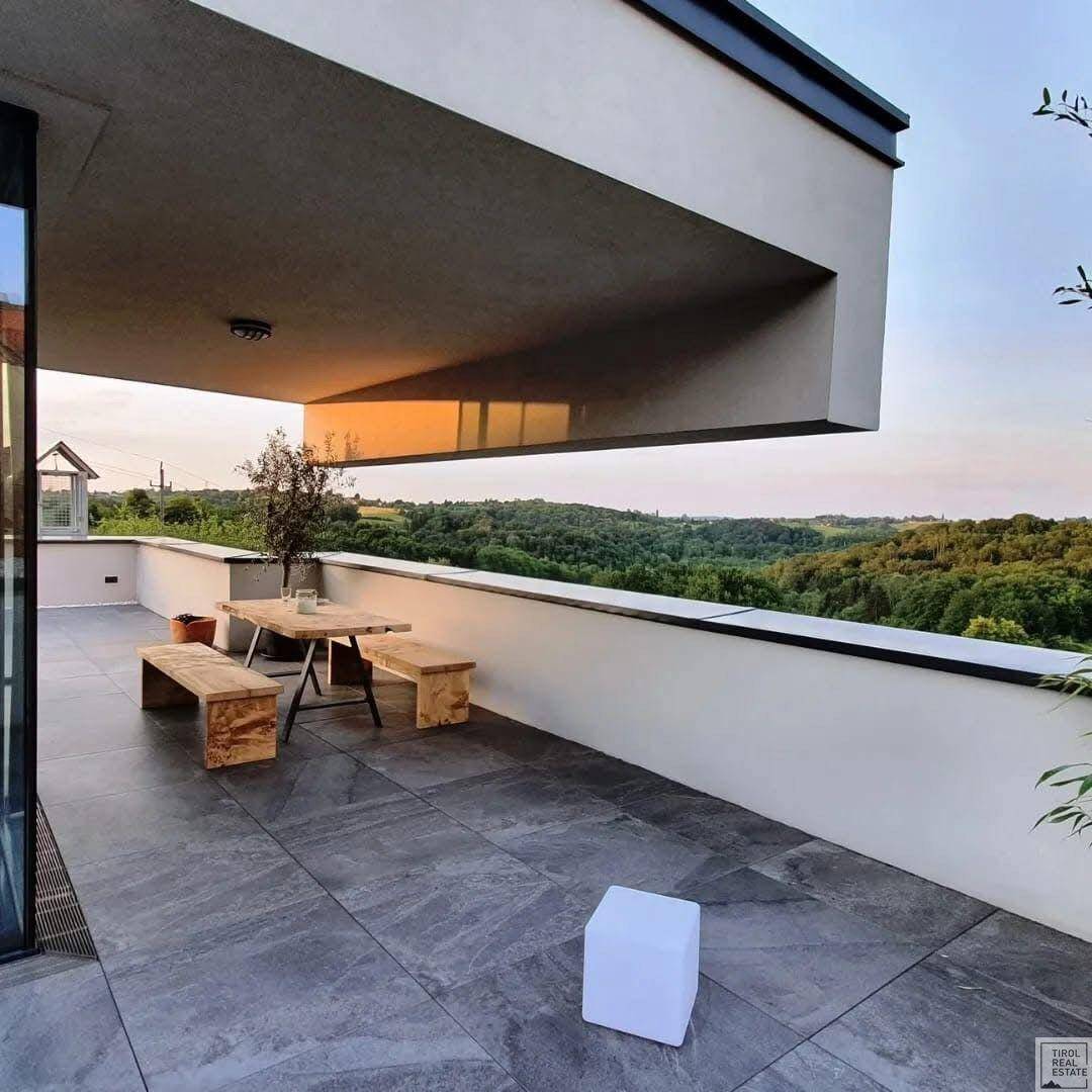 Geräumige Terrasse mit modernen Fliesen, Holztisch und Bänken, umgeben von grüner Landschaft.