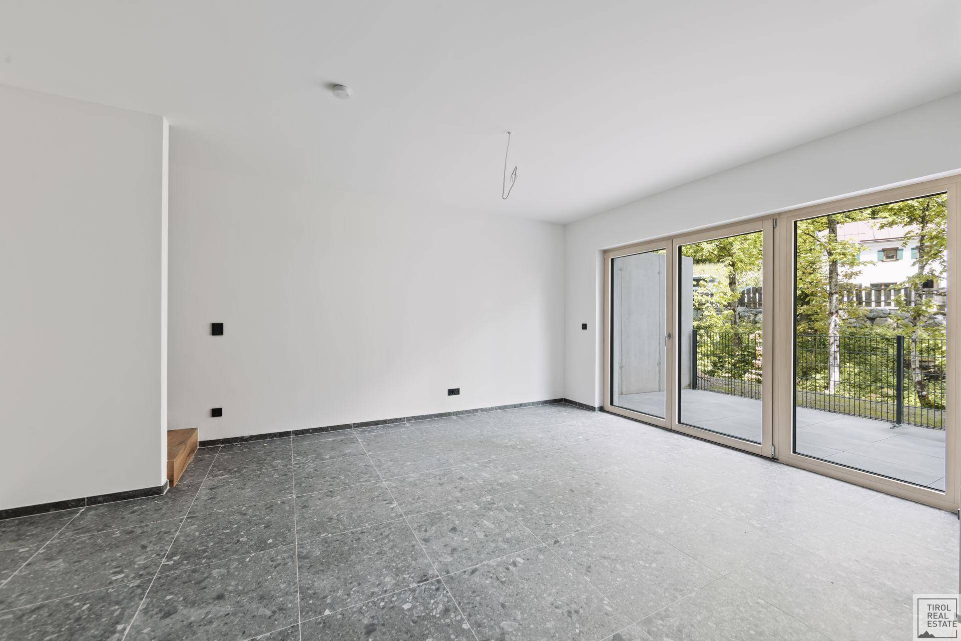 Heller Wohnbereich mit modernen grauen Fliesen und Zugang zu einem Außenbereich durch große Fenster.