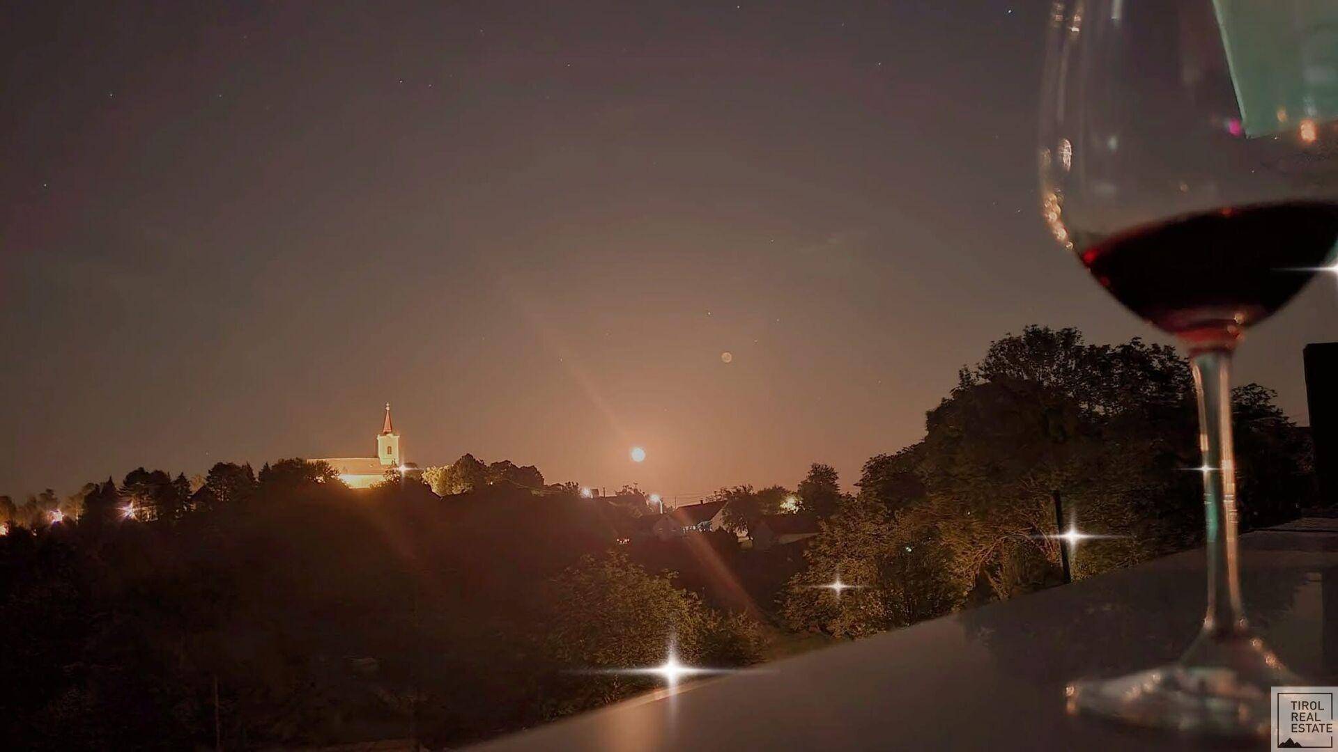 Weinglas auf einer Terrasse bei Nacht mit Blick auf ein beleuchtetes Dorf und den Mond.