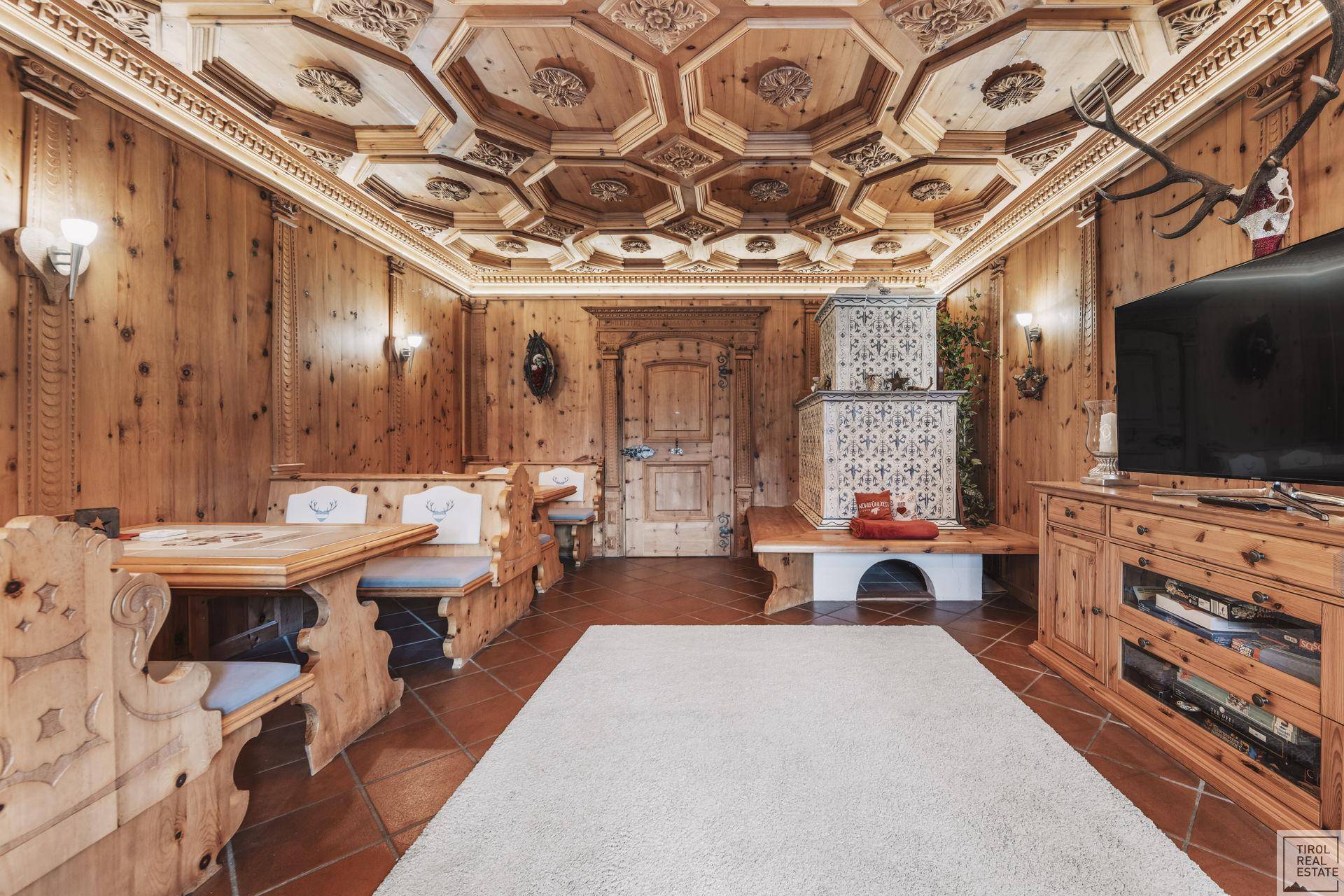 Traditionelle Stube mit aufwendig geschnitzter Holzdecke, Kachelofen und rustikalen Möbeln.