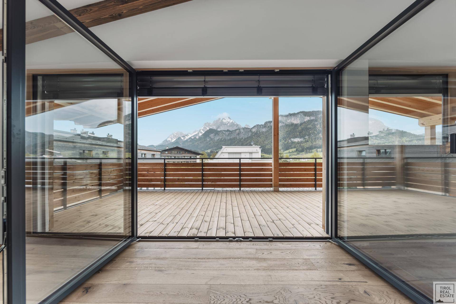 Blick von innen auf eine große Holzterrasse mit Bergblick durch eine breite Glasfront.