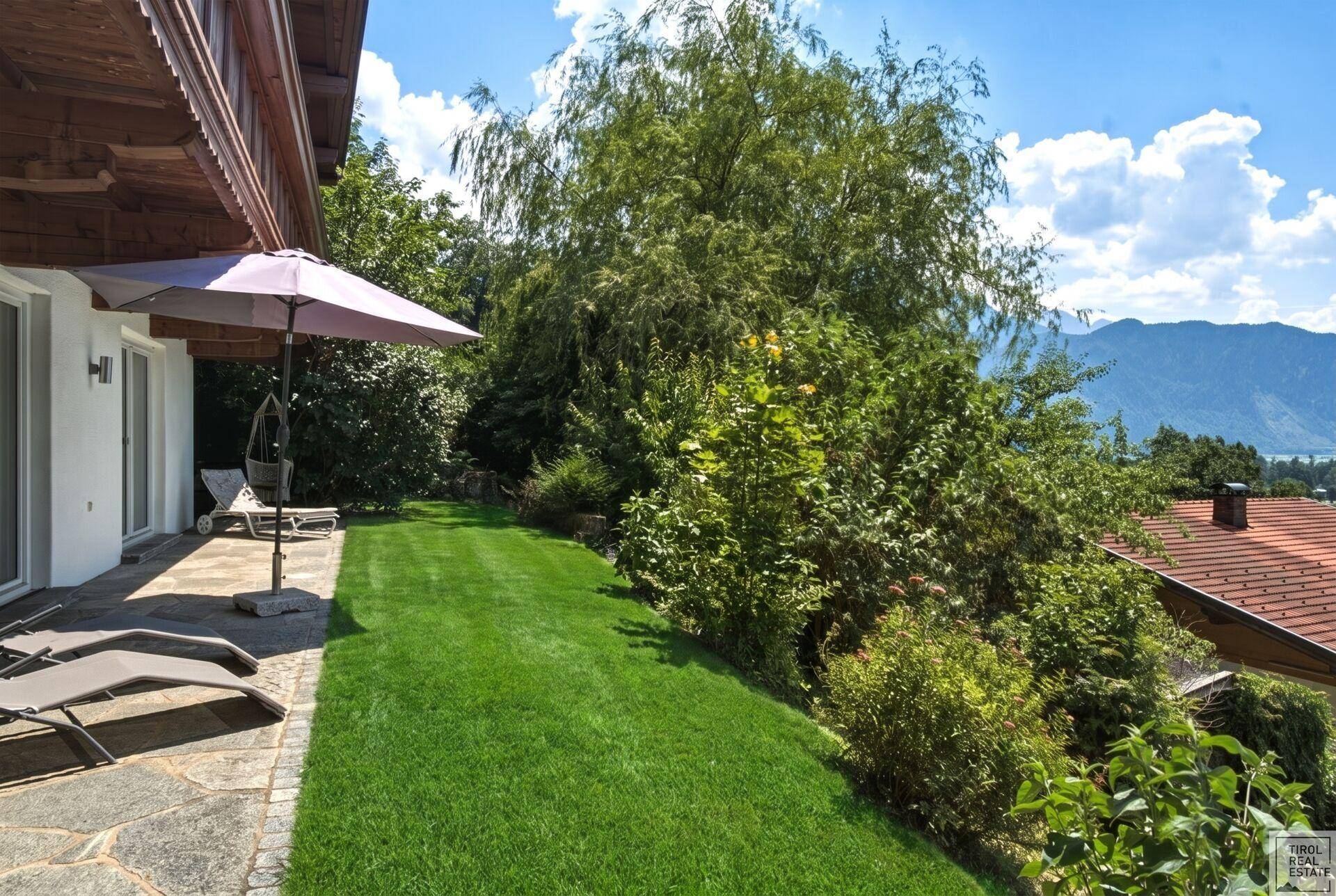 Gepflegter Garten mit weitläufigem Rasen, üppigen Bäumen und einem sonnigen Terrassenbereich mit Liegestühlen.