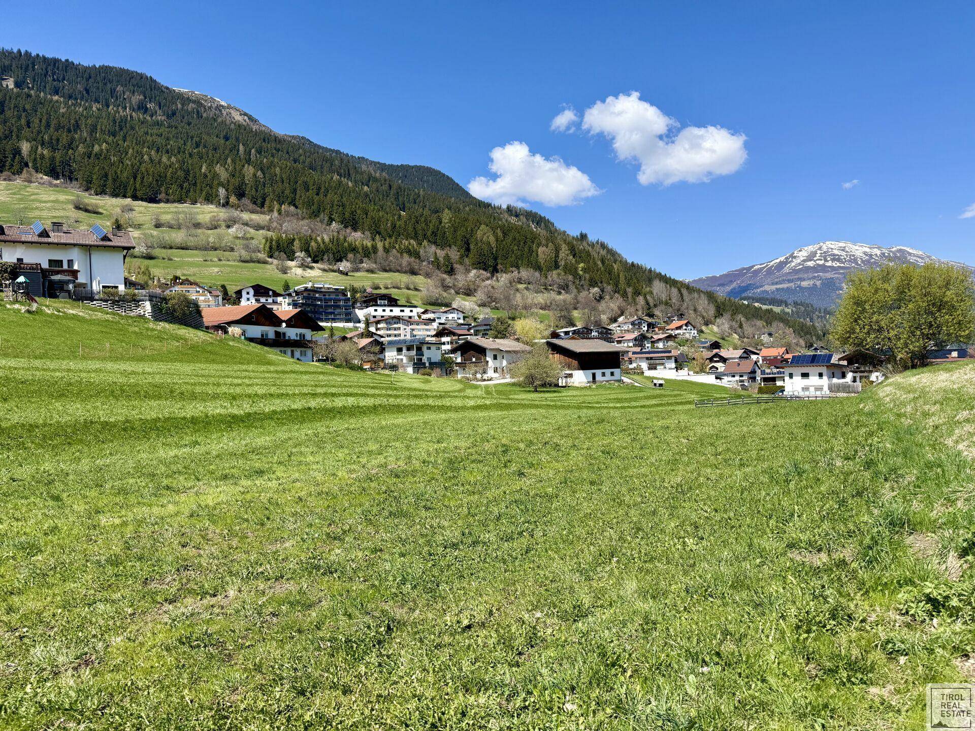 Panoramablick über saftige grüne Felder auf ein malerisches Dorf und die majestätischen Berge im Hintergrund.