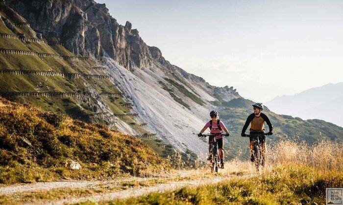 Zwei Personen beim Mountainbiken auf einem Bergpfad mit beeindruckender alpiner Kulisse im Hintergrund.