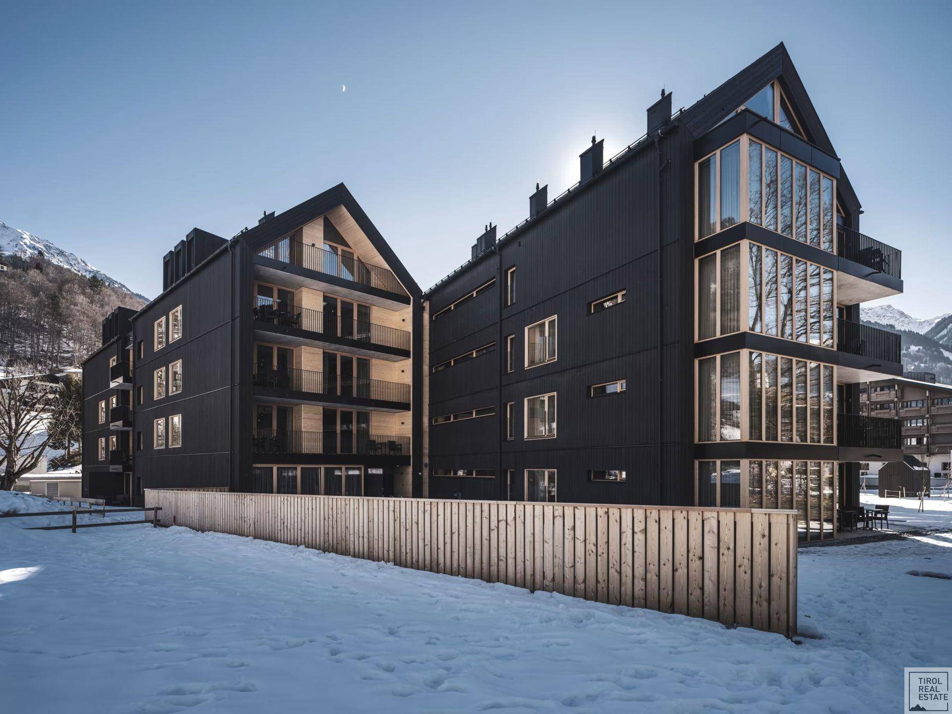 Moderne Architektur mit dunkler Fassade und großen Glasfronten in verschneiter Berglandschaft.