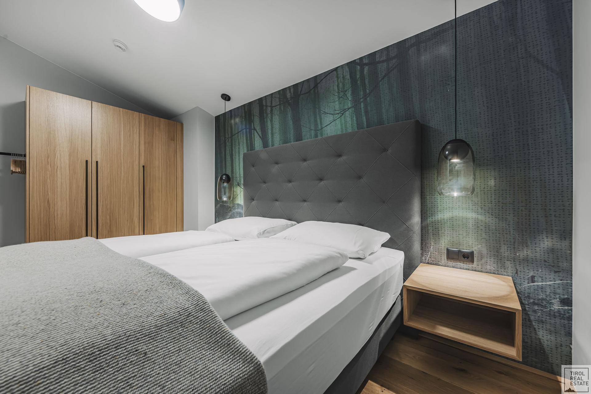 Schlafzimmer mit großem Doppelbett, Holzkleiderschrank und dunkler Wandtapete mit Waldmotiv.