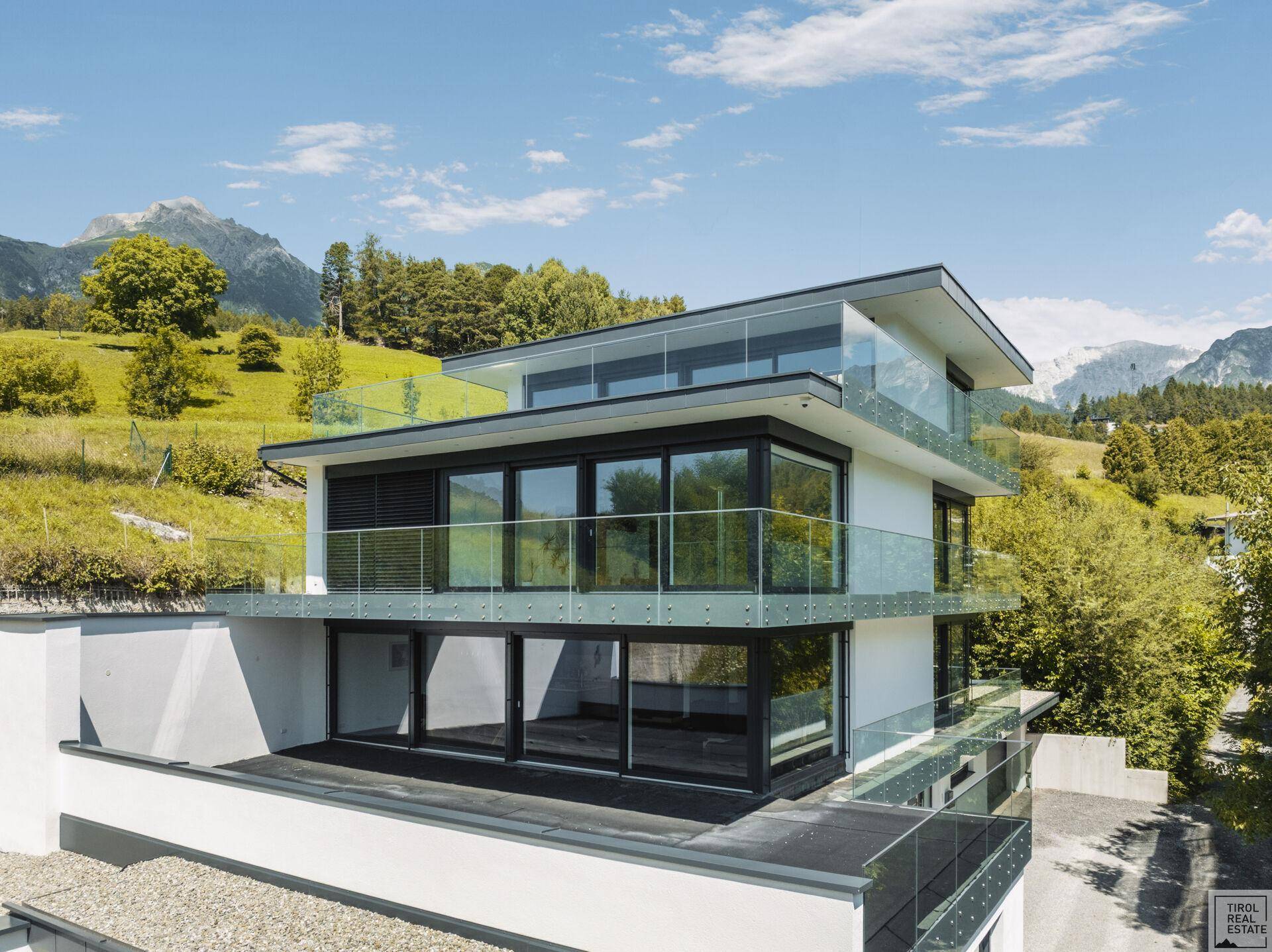 Elegante Fassade der modernen Villa mit großen Glasfronten und Balkonen vor alpiner Kulisse.