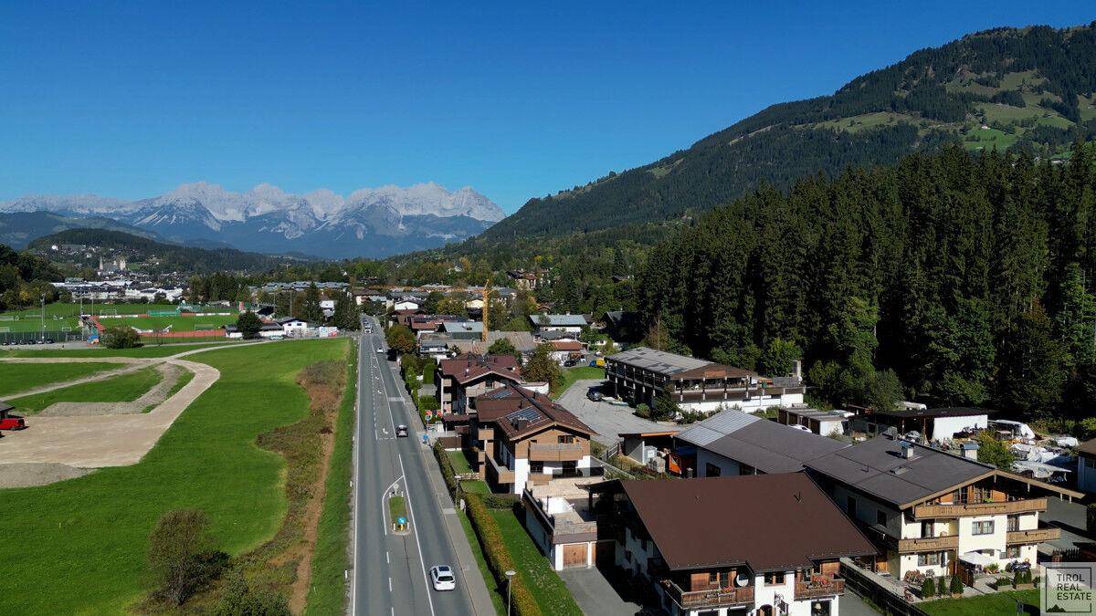 Panoramablick auf eine Siedlung entlang einer Straße, umgeben von Wäldern und einer weiten Bergkette am Horizont.