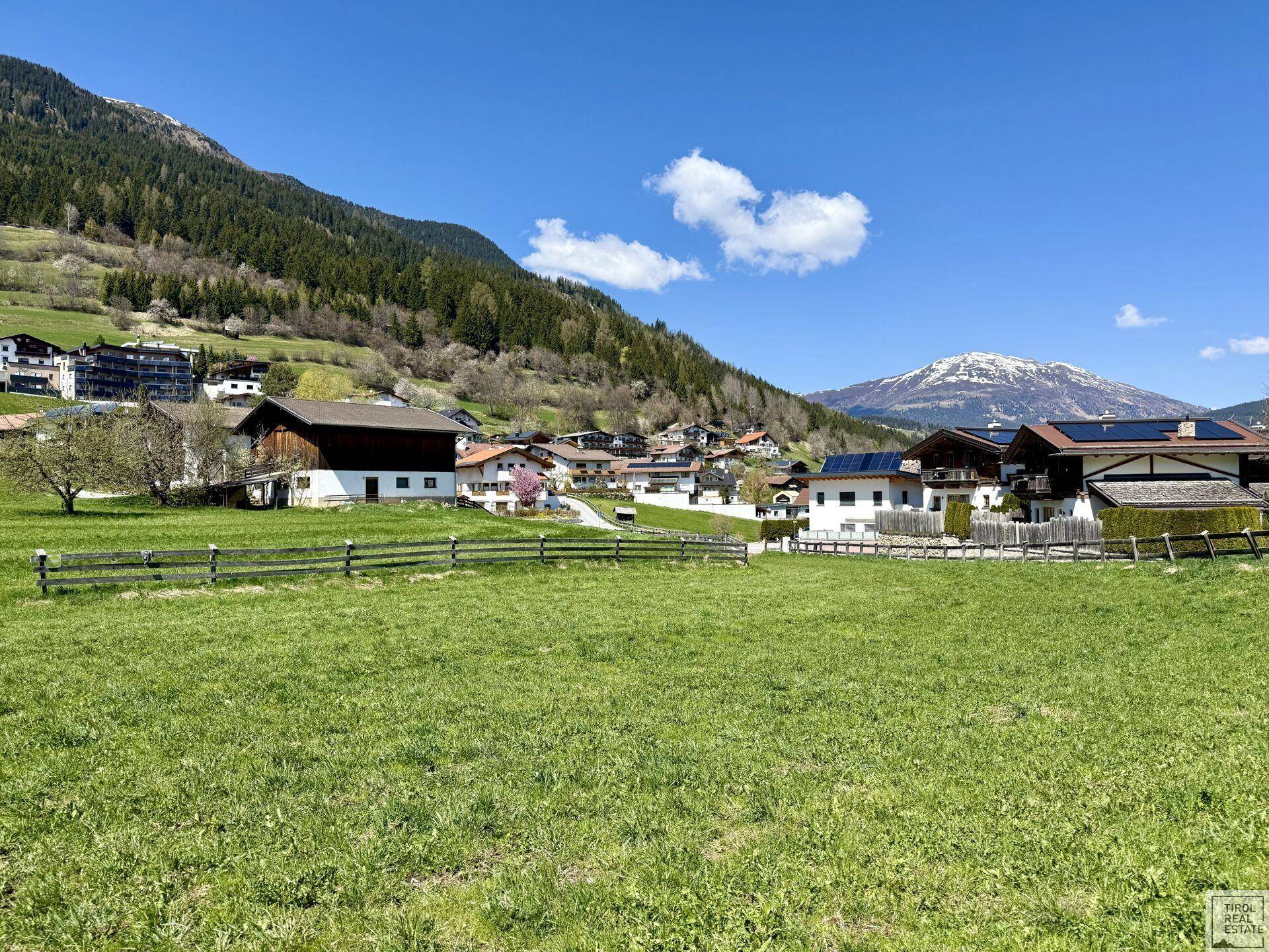 Sonnige Landschaft mit grünen Wiesen, einem traditionellen Dorf und schneebedeckten Berggipfeln am Horizont.