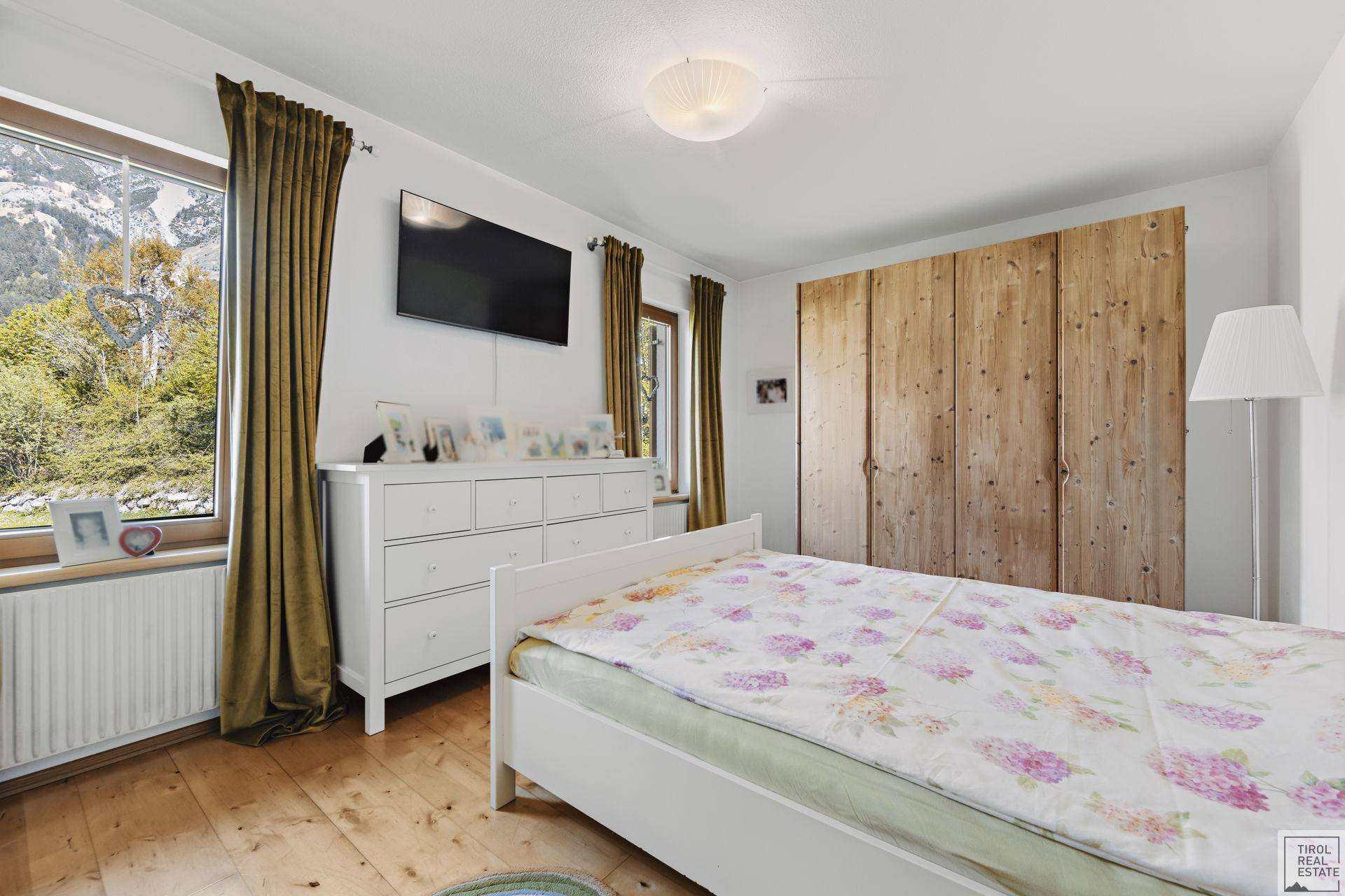 Helles Schlafzimmer mit weißem Bett, Holzschrank und Blick ins Grüne durch das Fenster.