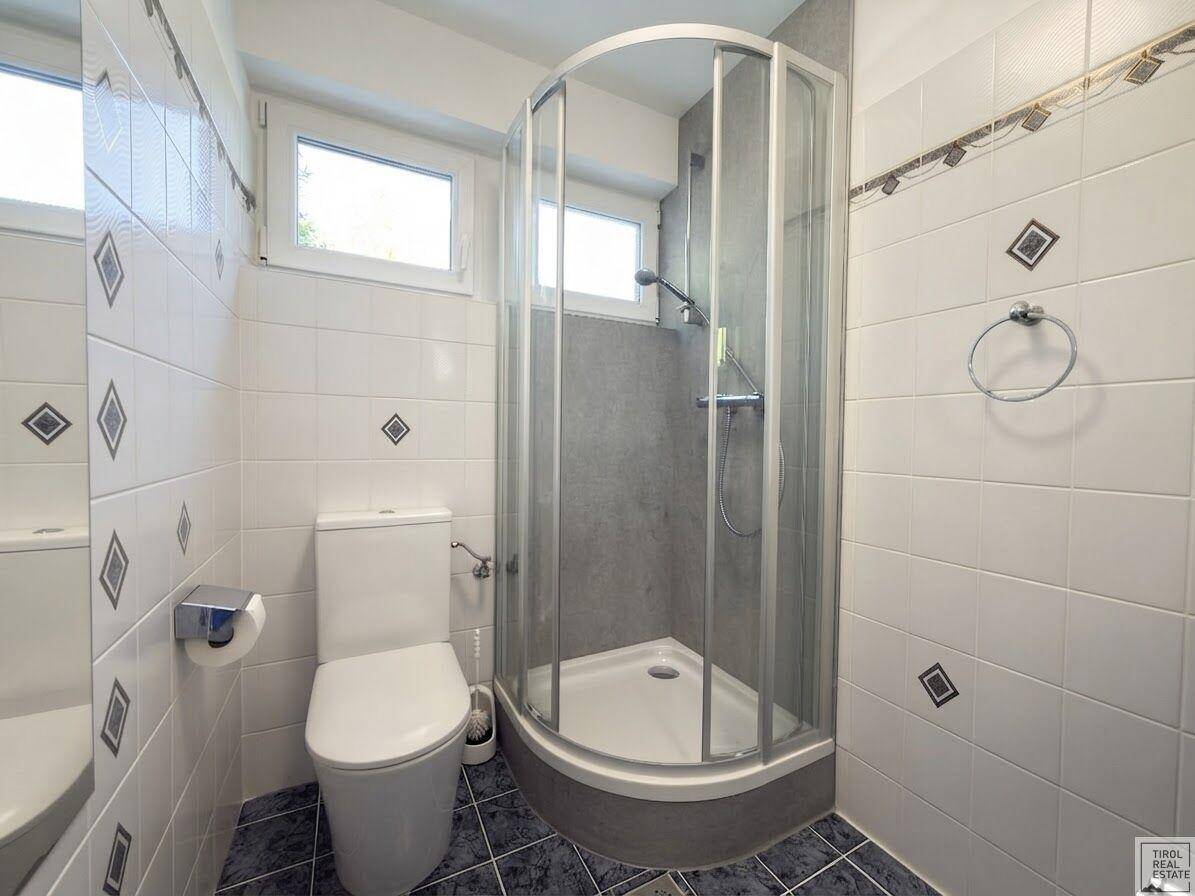 Gefliestes Badezimmer mit Duschkabine und Toilette, ausgestattet mit zwei Fenstern.