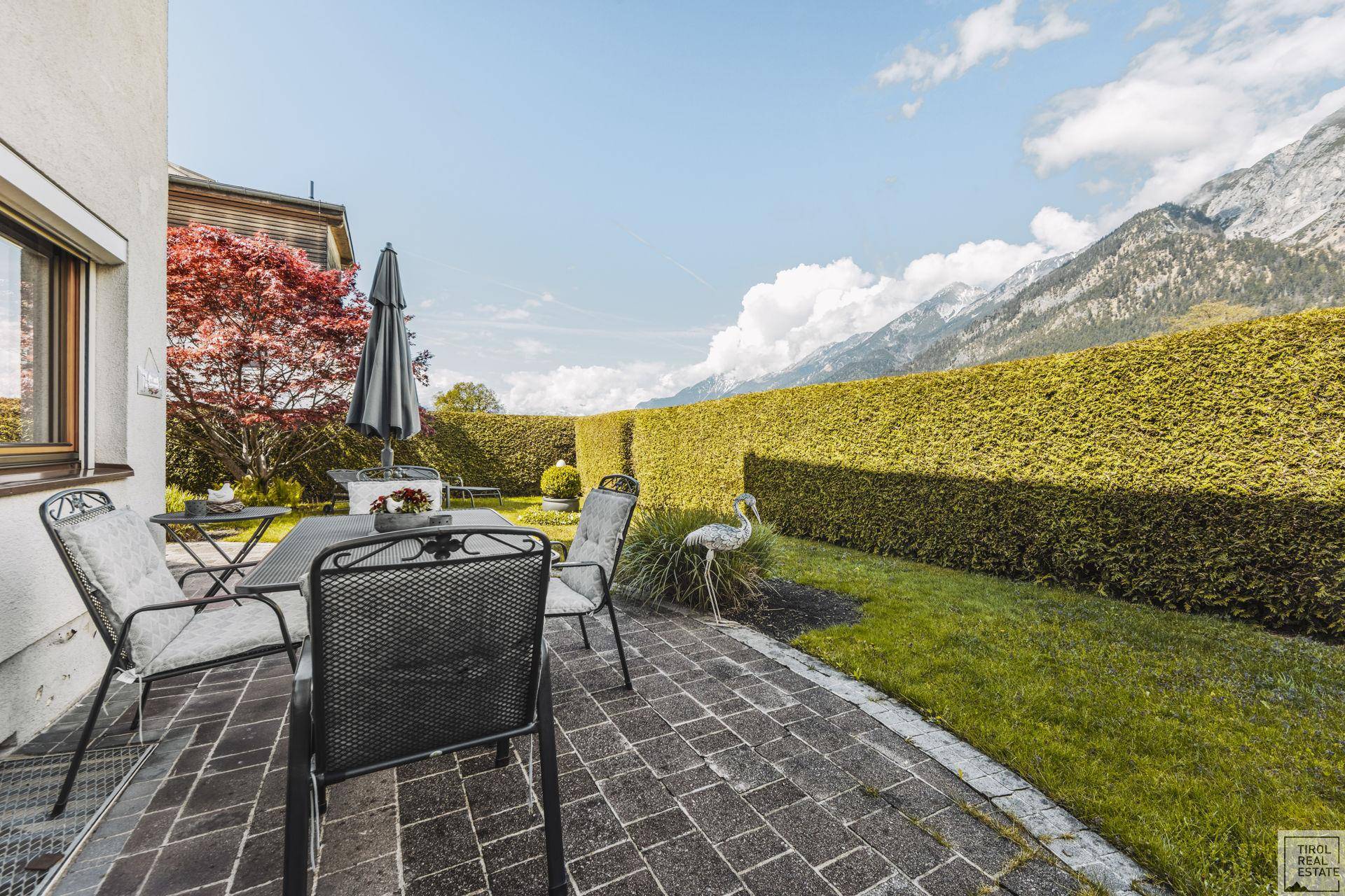 Gemütliche Terrasse mit Esstisch, Stühlen und einer hohen Hecke vor der Bergkulisse.