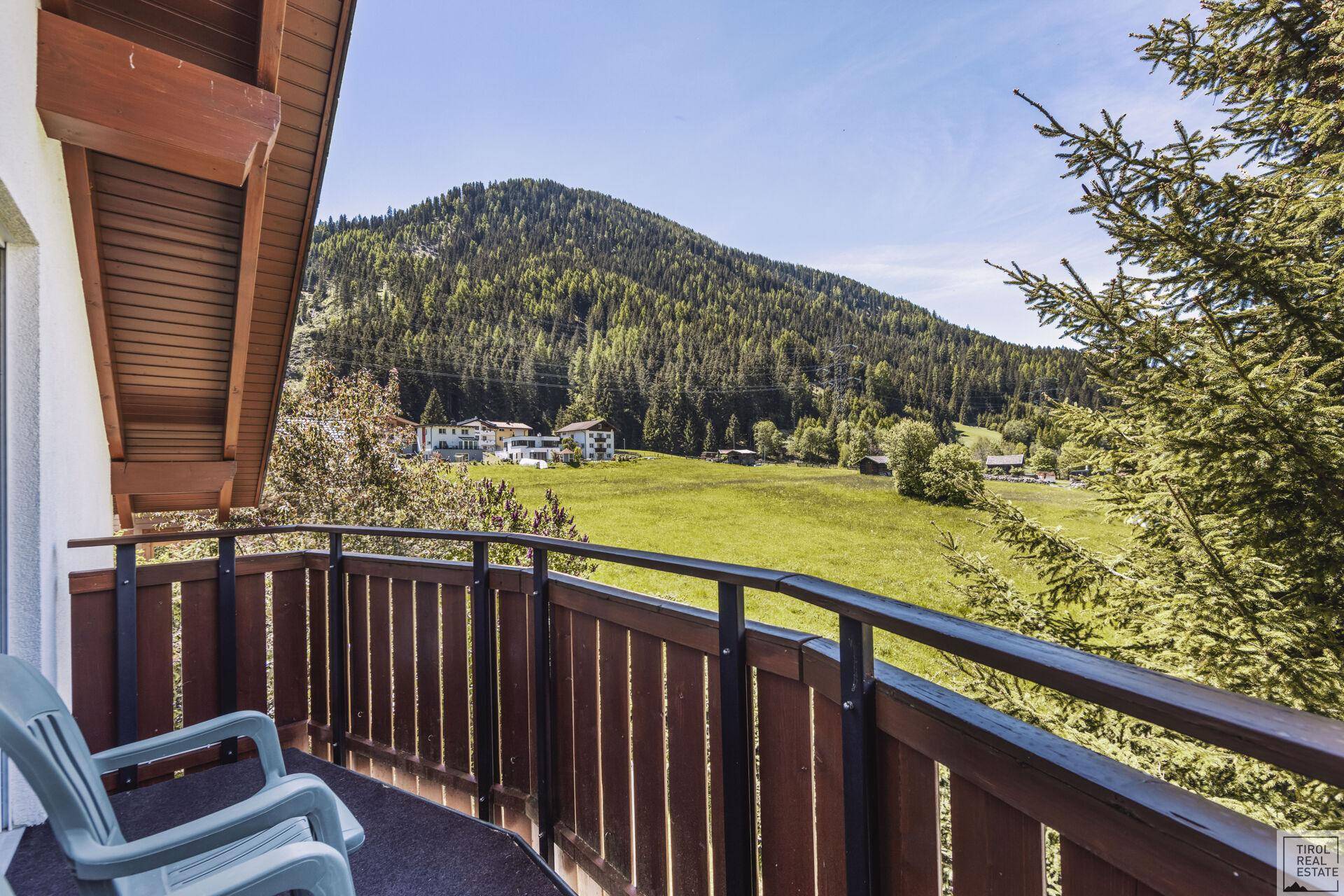 Balkon mit Blick auf grüne Wiesen und bewaldete Hügel unter blauem Himmel.