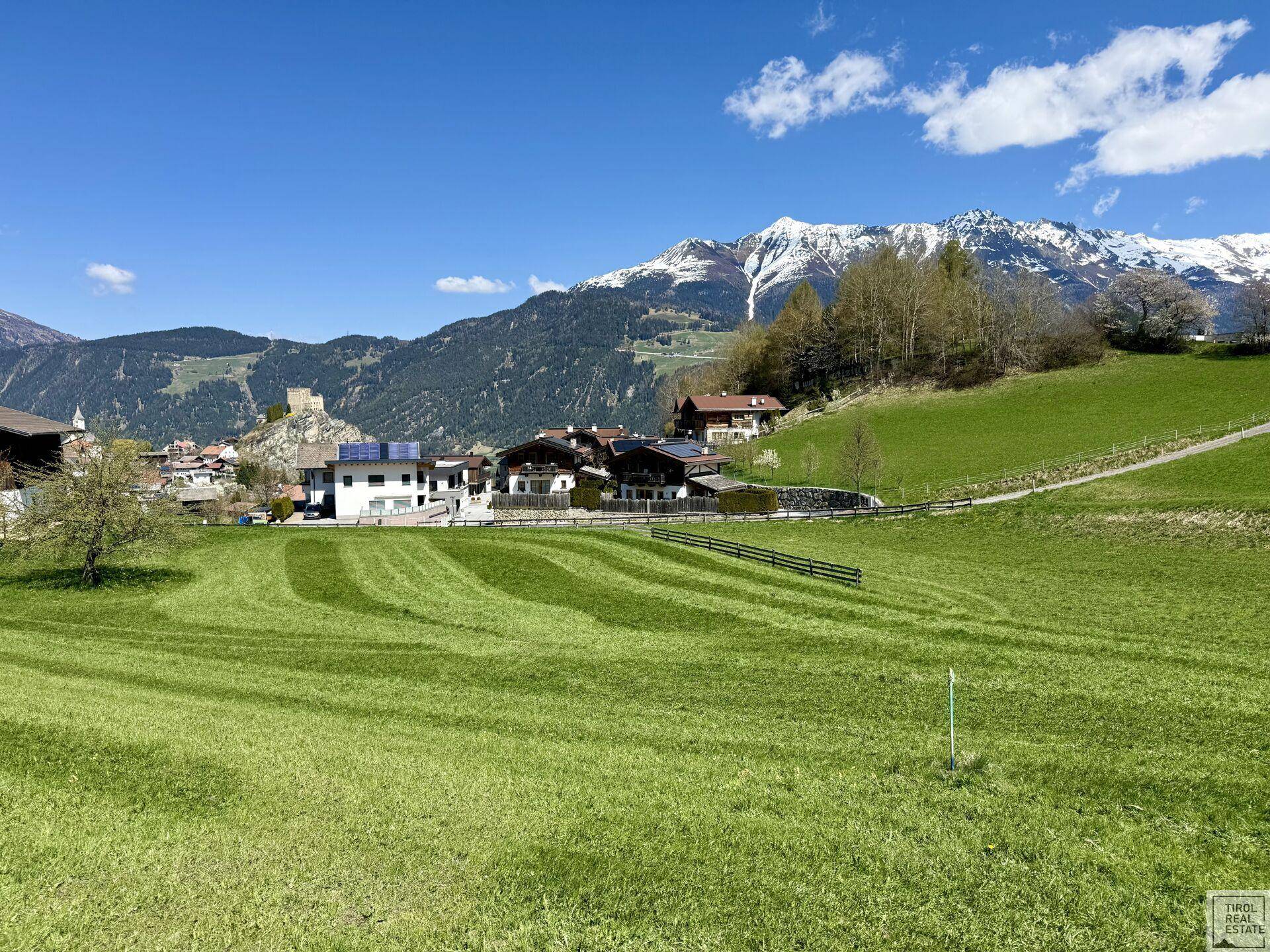 Grüne Felder mit Blick auf ein charmantes Dorf, eine historische Burgruine und die imposante Bergkulisse.