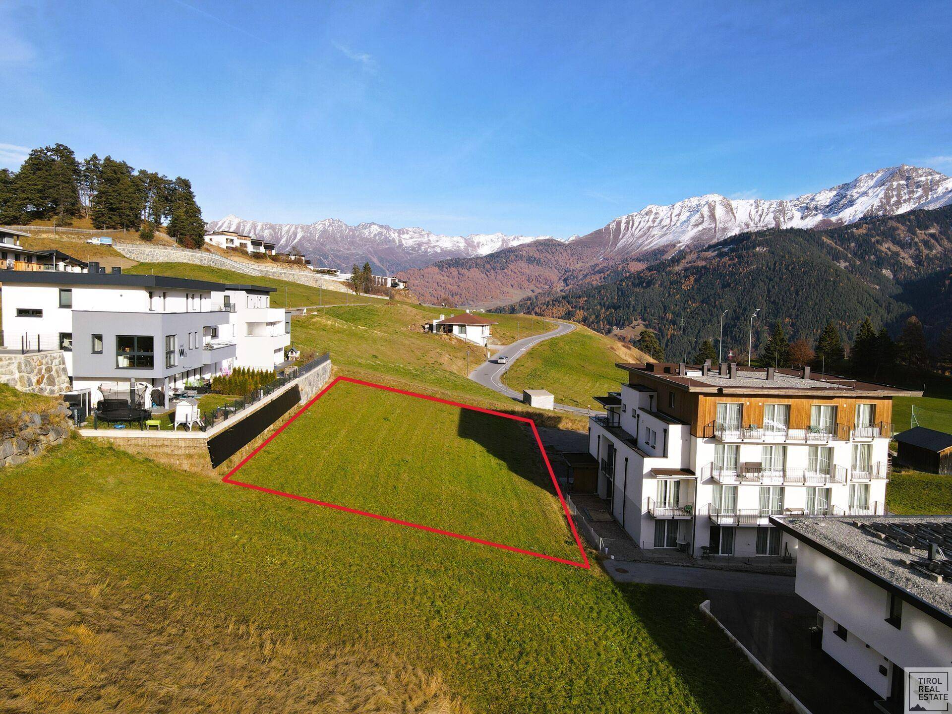 Sonniges Baugrundstück in alpiner Umgebung, umgeben von modernen Gebäuden und mit Bergblick.