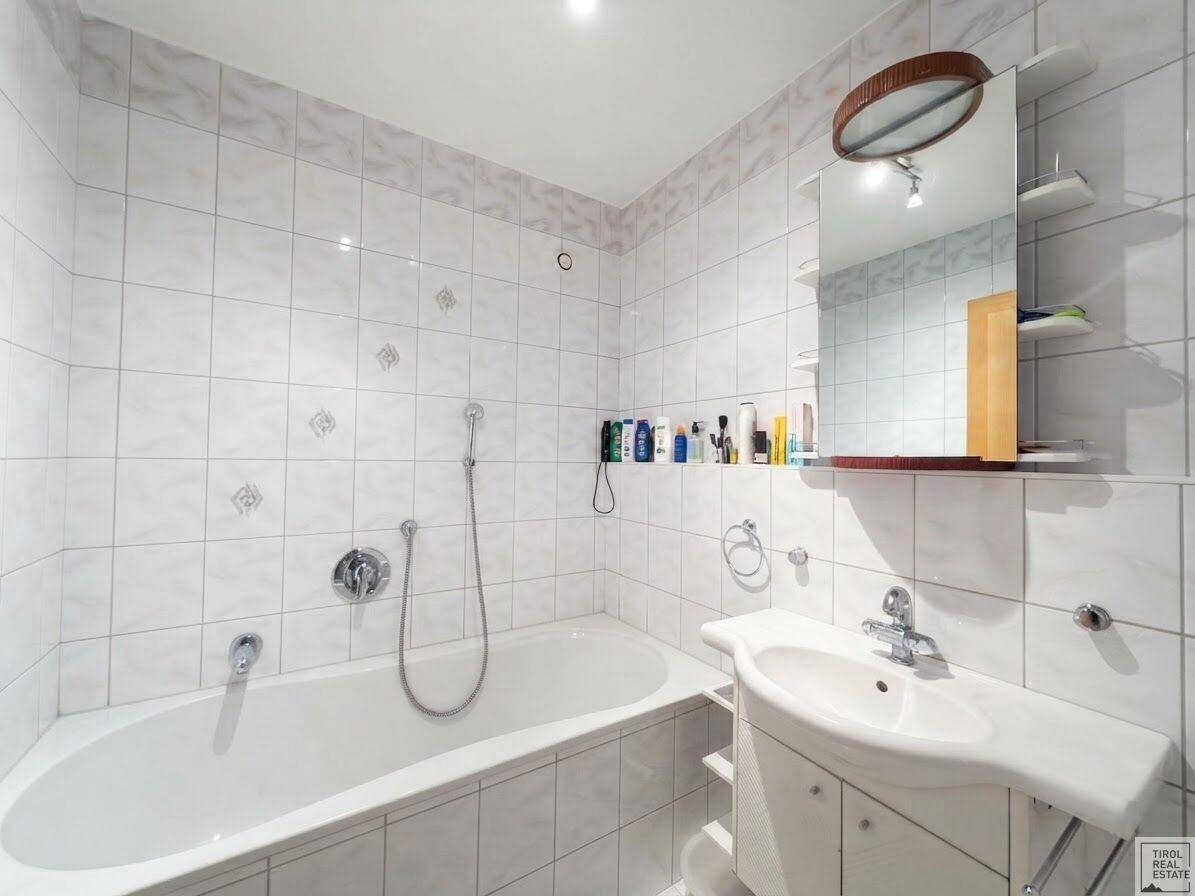 Helles Badezimmer mit weißen Fliesen, Badewanne mit Duschfunktion und Einzelwaschbecken.