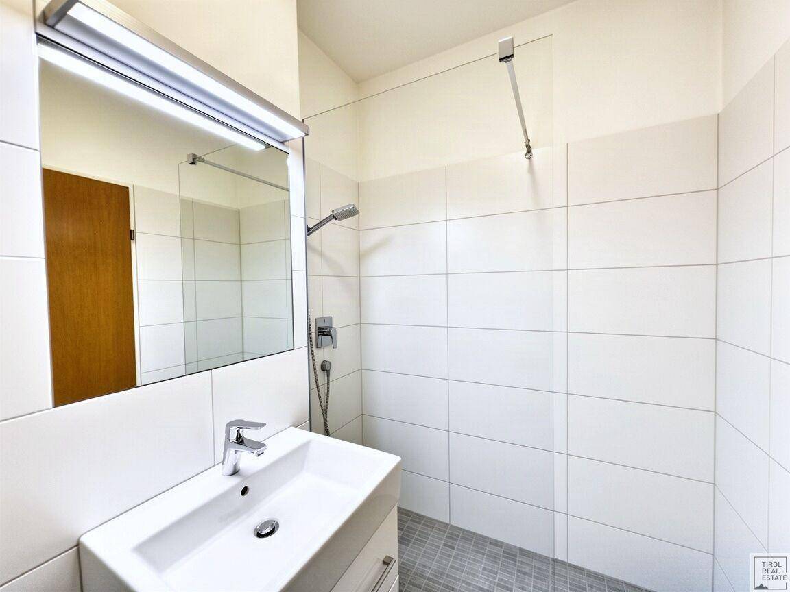Modernes Badezimmer mit weißem Waschbecken, Spiegel und einer geräumigen, gefliesten begehbaren Dusche.